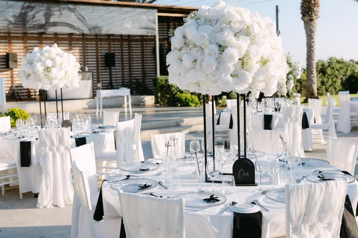 Monochrome wedding reception styling with tall white rose globe centrepieces on slim black stands, beaded charger plates, black napkins and acrylic table signage on round tables