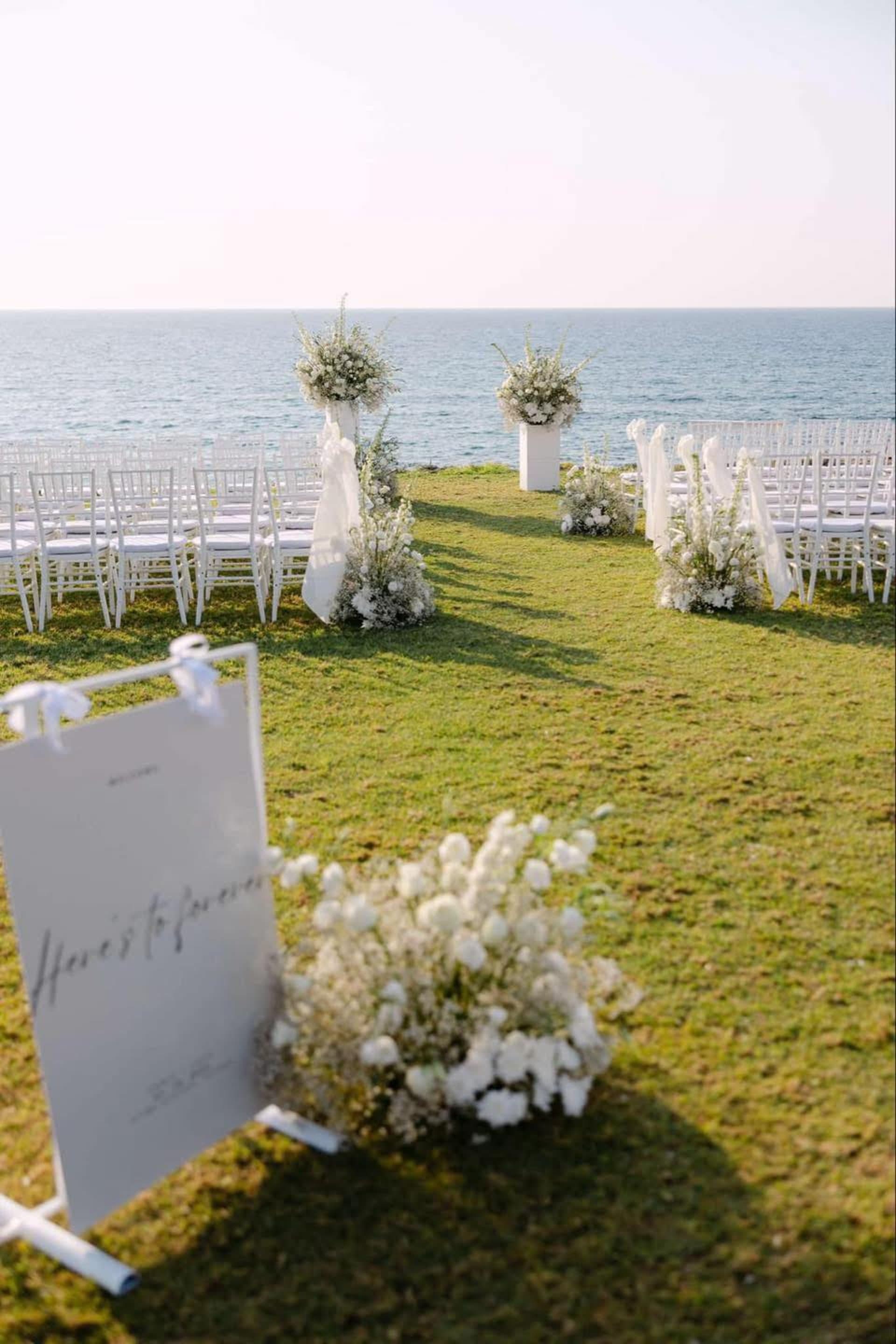 All-white wedding ceremony setup on the clifftop lawn at Alassos, with white chiavari chairs, draped aisle markers, tall white floral arrangements on plinths and a calligraphy welcome sign, facing the open sea