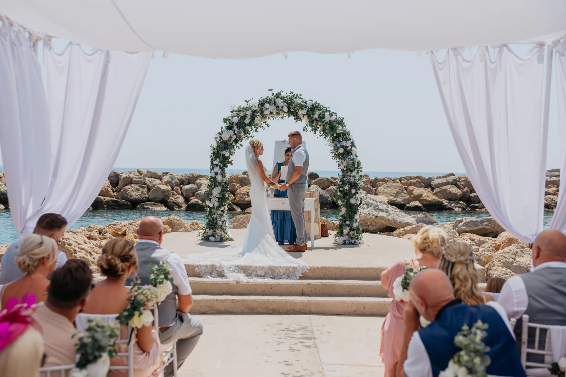 Guests seated under white drapes as a couple marry at the seafront arch, Coral Beach