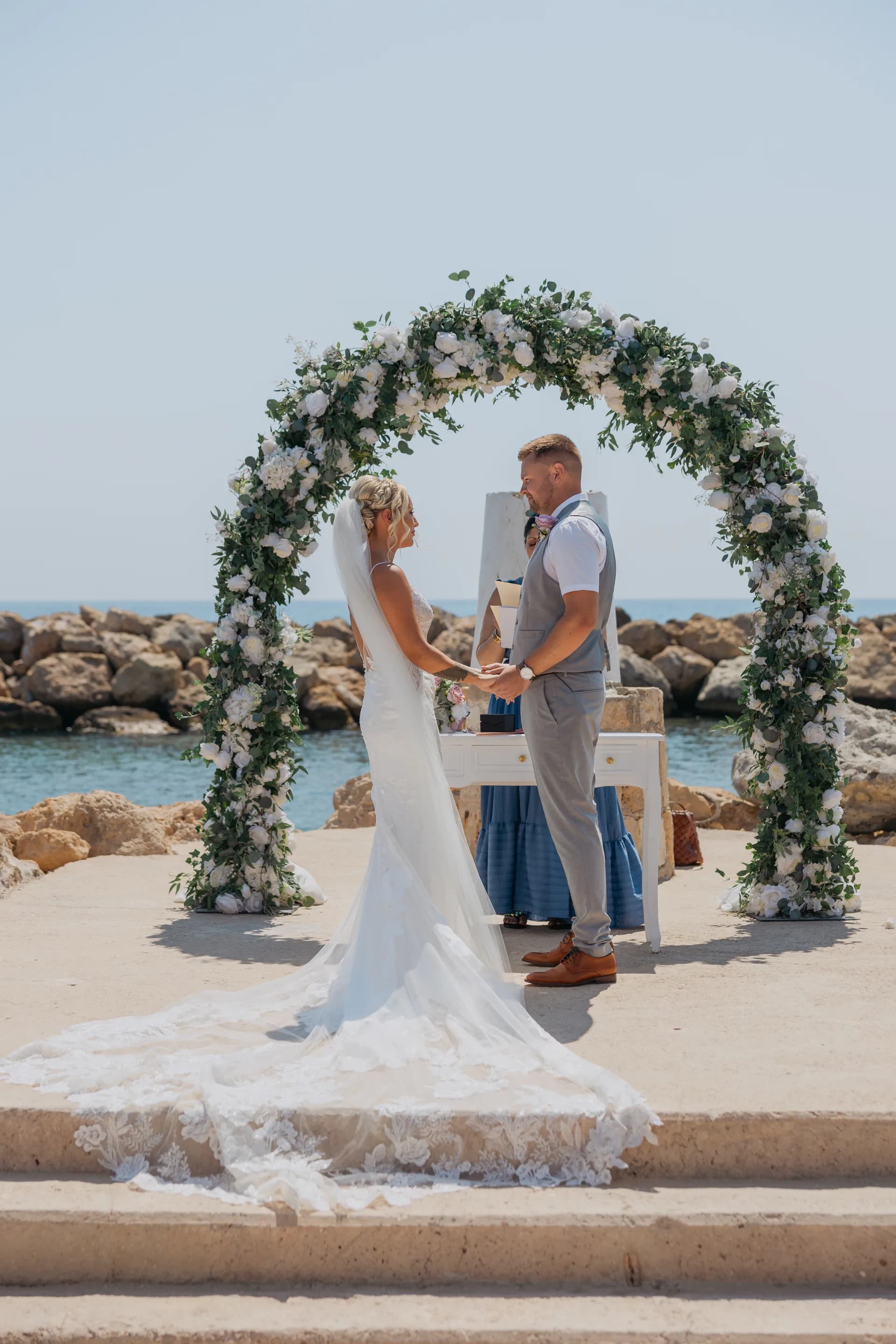 Bride and groom under the white and green floral arch on the rocky point at Coral Beach