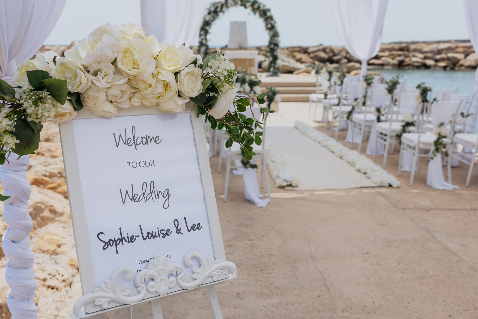 Welcome sign topped with white roses beside the ceremony aisle at Coral Beach, sea behind