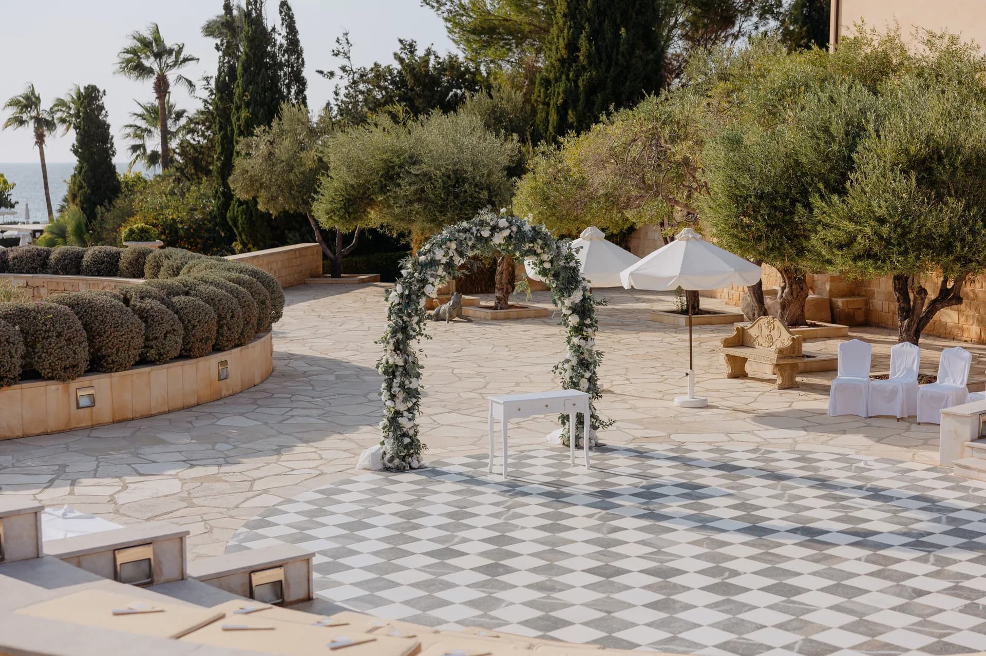 The Elysium’s dressed floral ceremony arch on the chequerboard terrace, with olive trees, parasols and a sliver of sea
