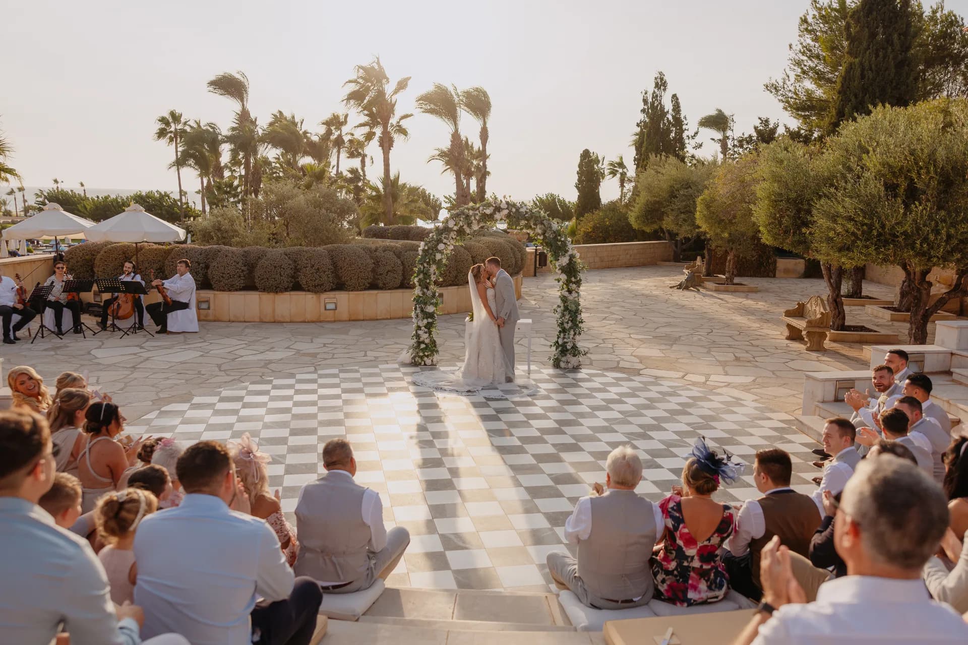 Backlit wide of the first kiss at the floral arch, guests around the Elysium’s chequerboard ceremony terrace