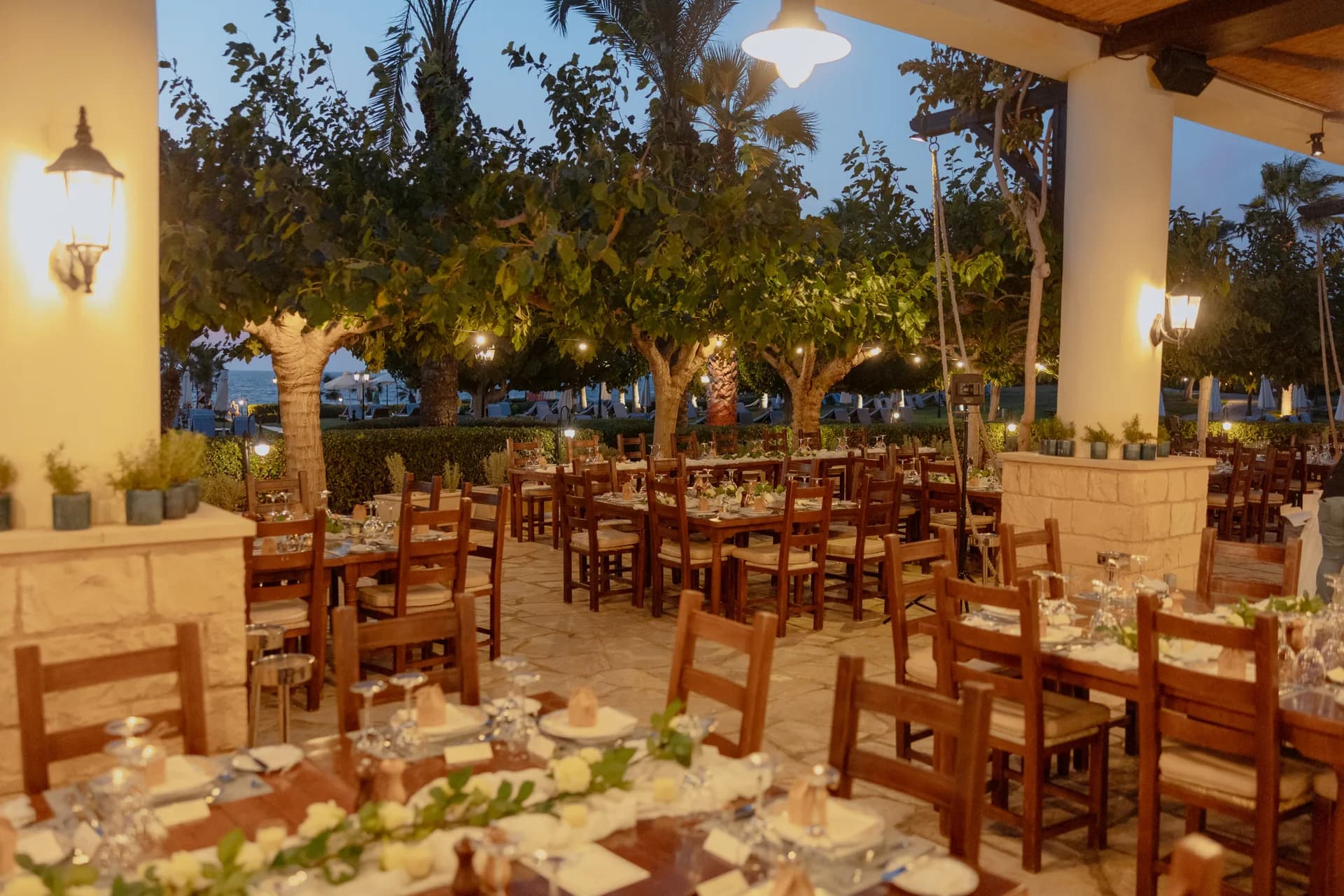 The Elysium’s covered dining terrace at blue hour, wall lanterns lit above laid tables with the sea beyond