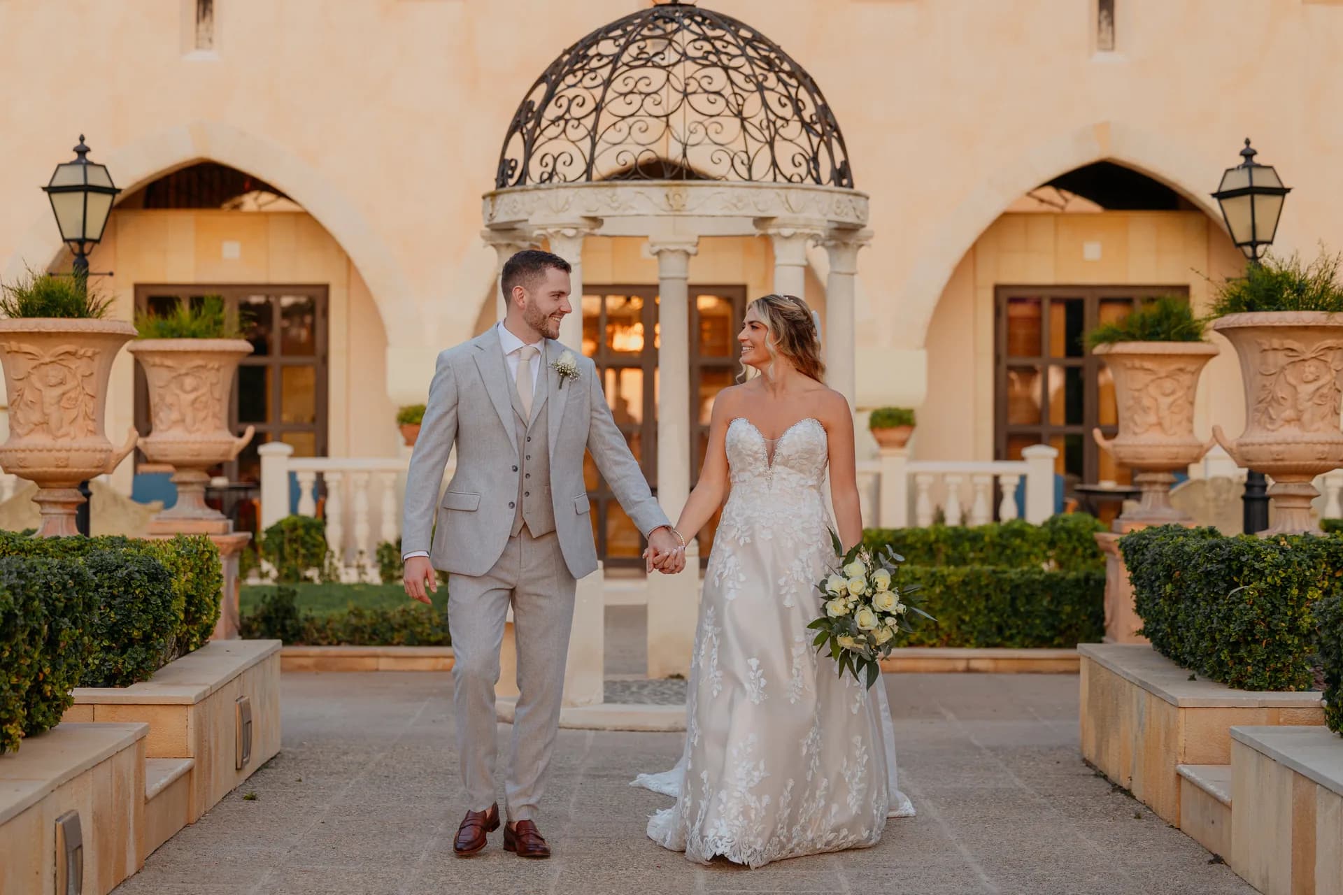 Bride and groom walk from the Elysium’s wrought-iron domed gazebo between carved stone urns at sunset