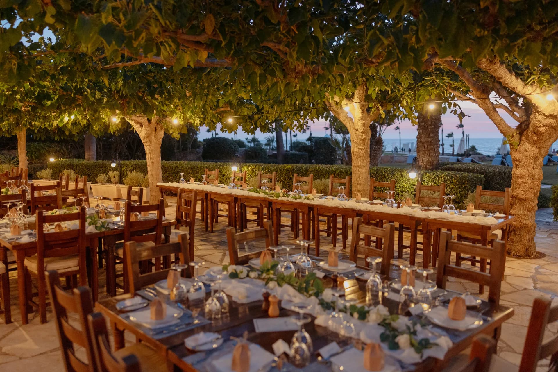 Long banquet tables set beneath festoon-lit trees at twilight, the sea glowing beyond the Elysium’s garden terrace