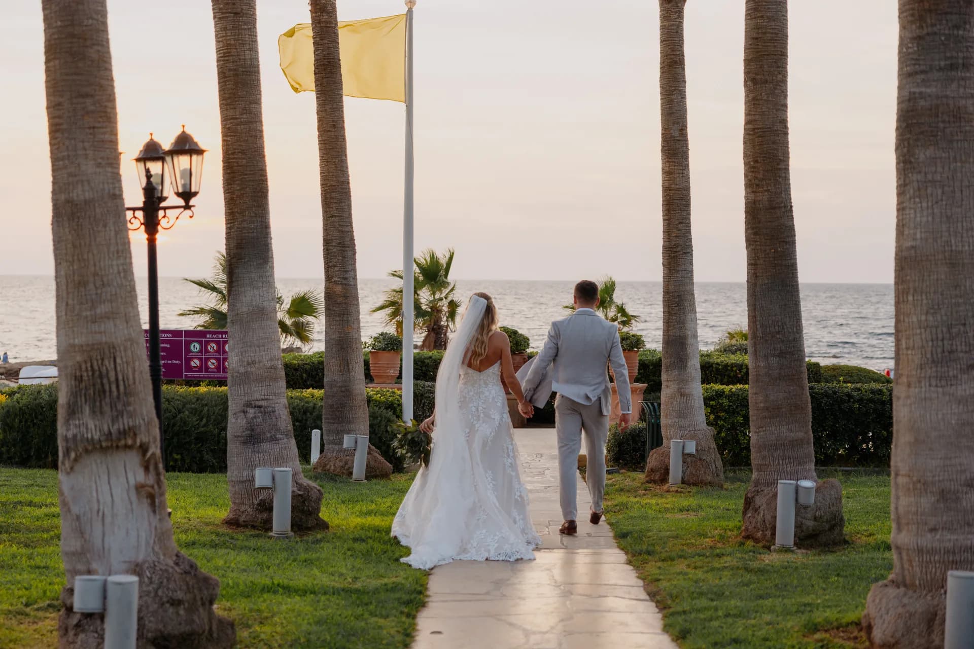 Bride and groom walking a palm-lined path across the Elysium’s lawns down to the Mediterranean