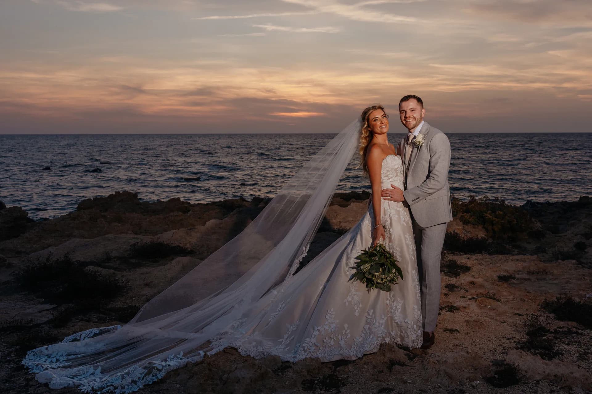 Bride and groom on the rocky shoreline below the Elysium at sunset, a cathedral veil sweeping across the frame