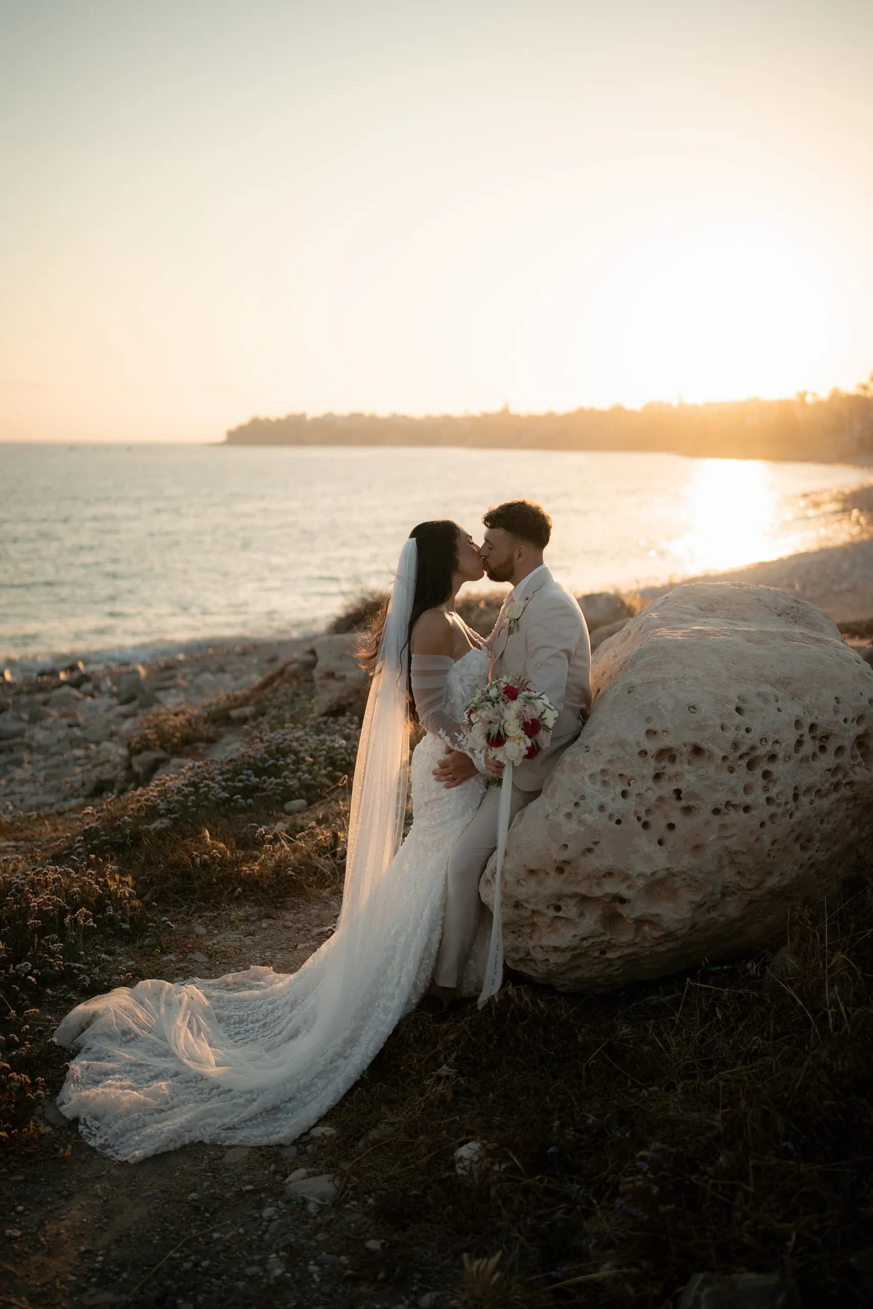 Newlyweds beside a pale sea rock at Coral Residence during a Cyprus sunset, the bride's veil and train sweeping across the stone