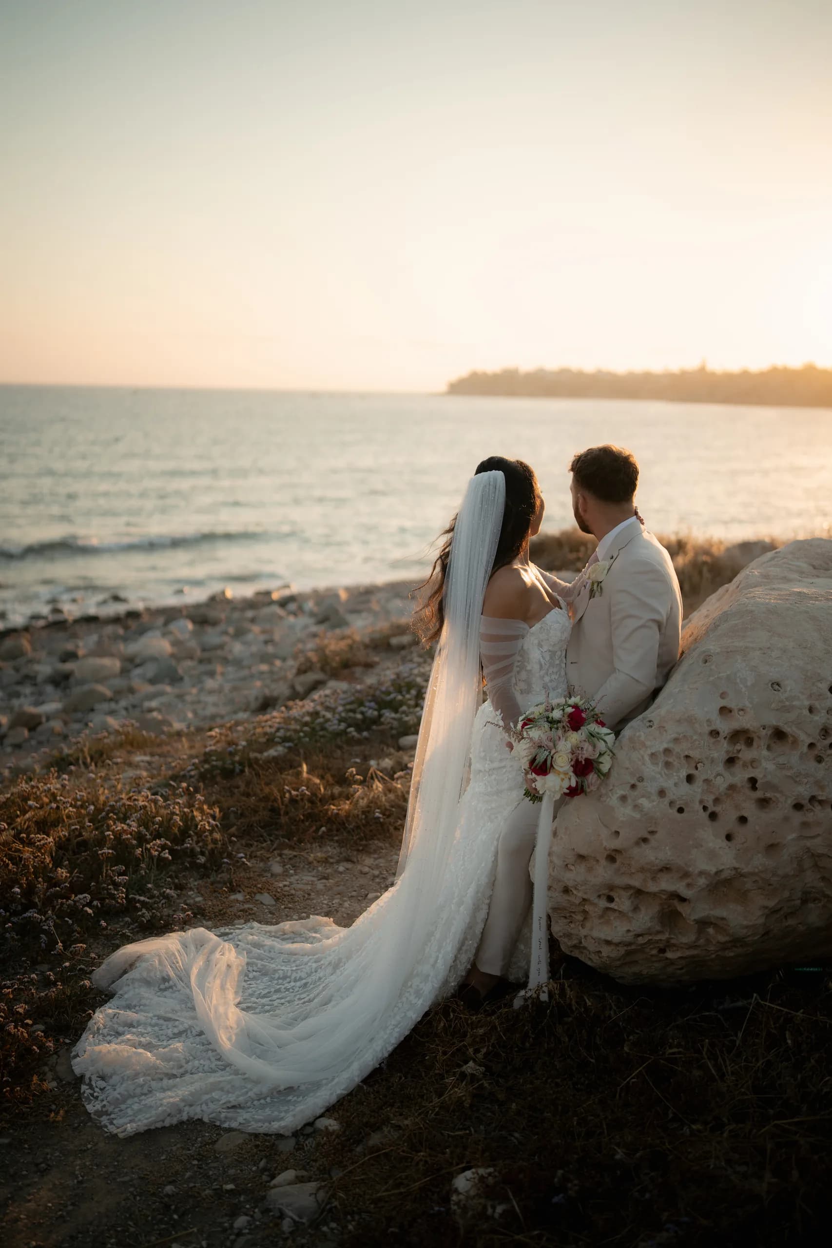 Bride and groom holding each other close at the water's edge at Coral Residence, golden sunlight low on the horizon behind them