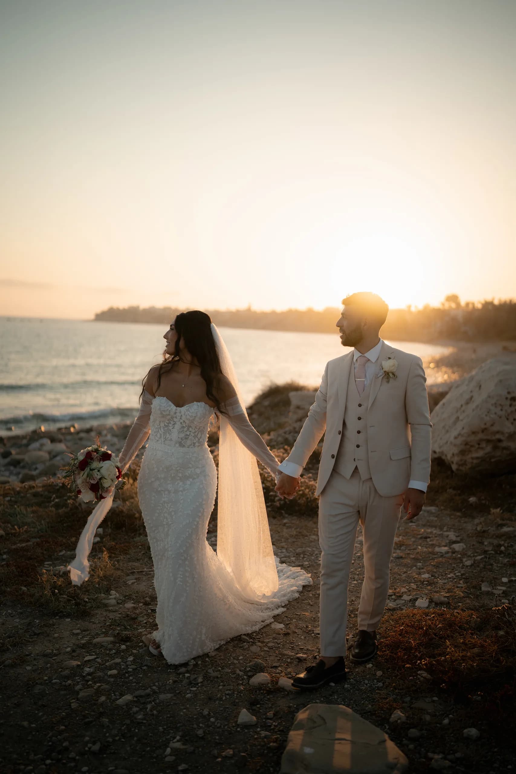 Bride and groom walking hand in hand toward the setting sun along the rocky shoreline at Coral Residence, Cyprus