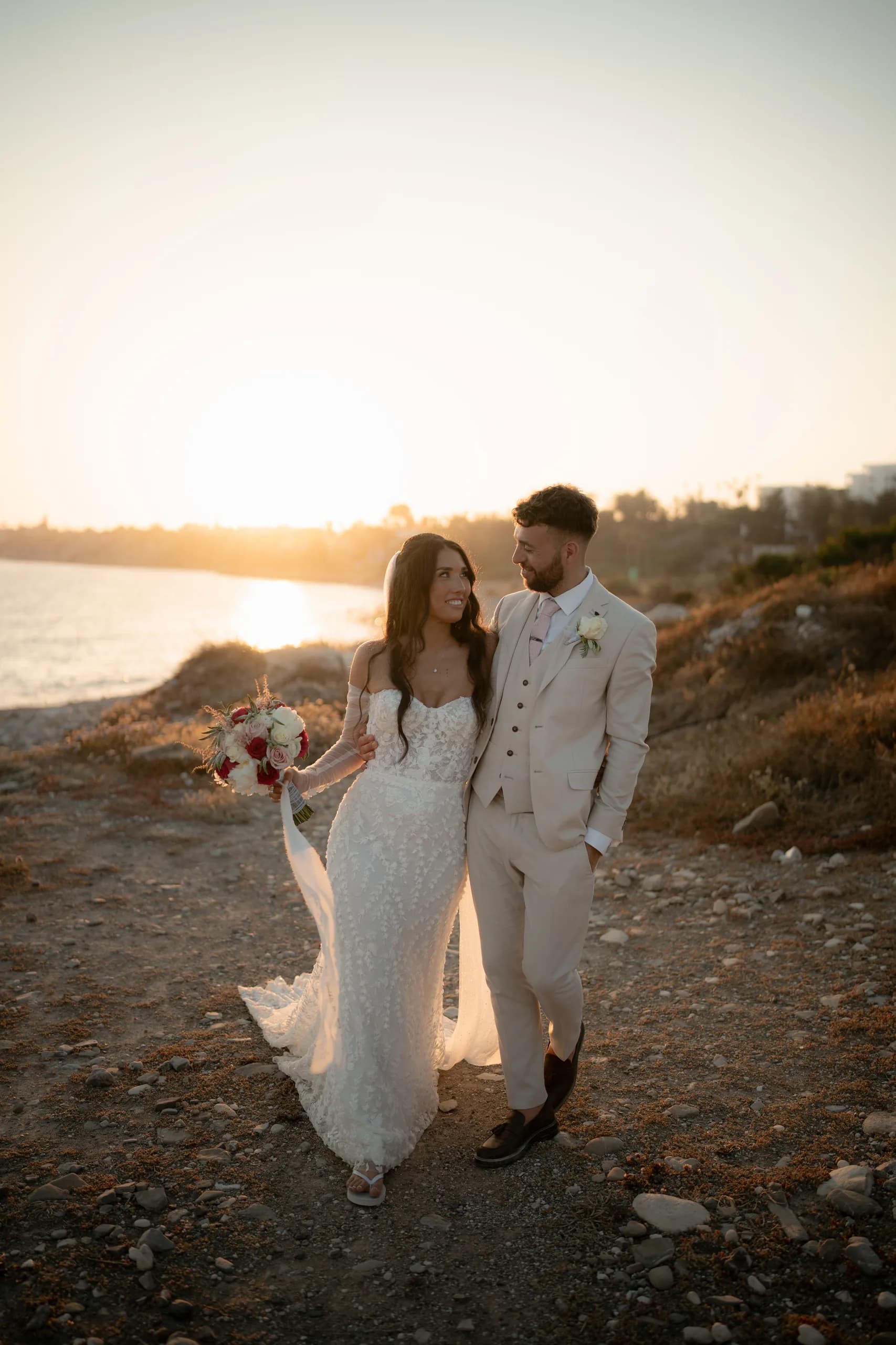 Bride holding her bouquet and groom walking back toward the camera at golden hour, the sun setting over the sea behind them at Coral Residence