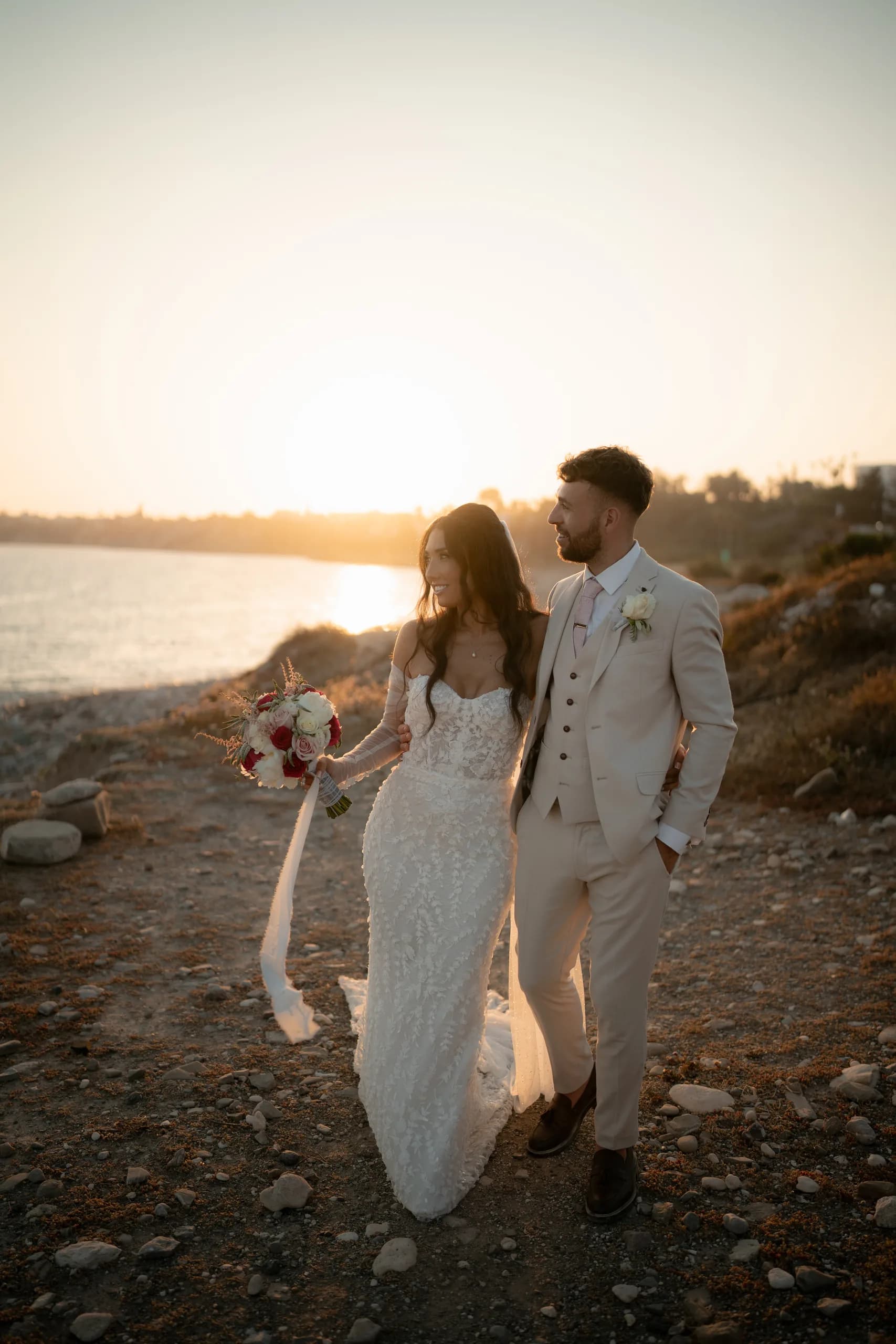 Newlyweds walking together along the coast at Coral Residence as the sun sinks toward the Mediterranean horizon