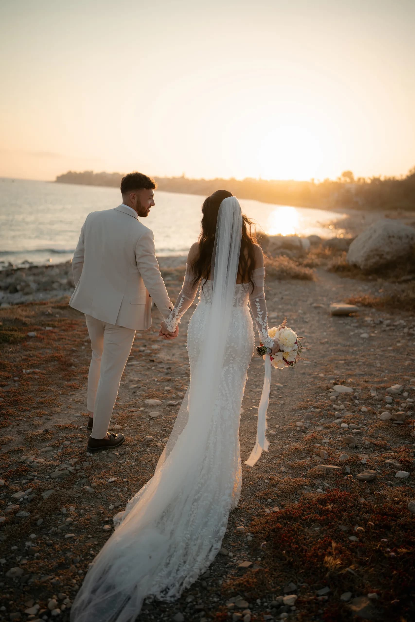 Bride and groom walking away toward the sunset at Coral Residence, the bride's veil and train trailing across the rocks in the golden light