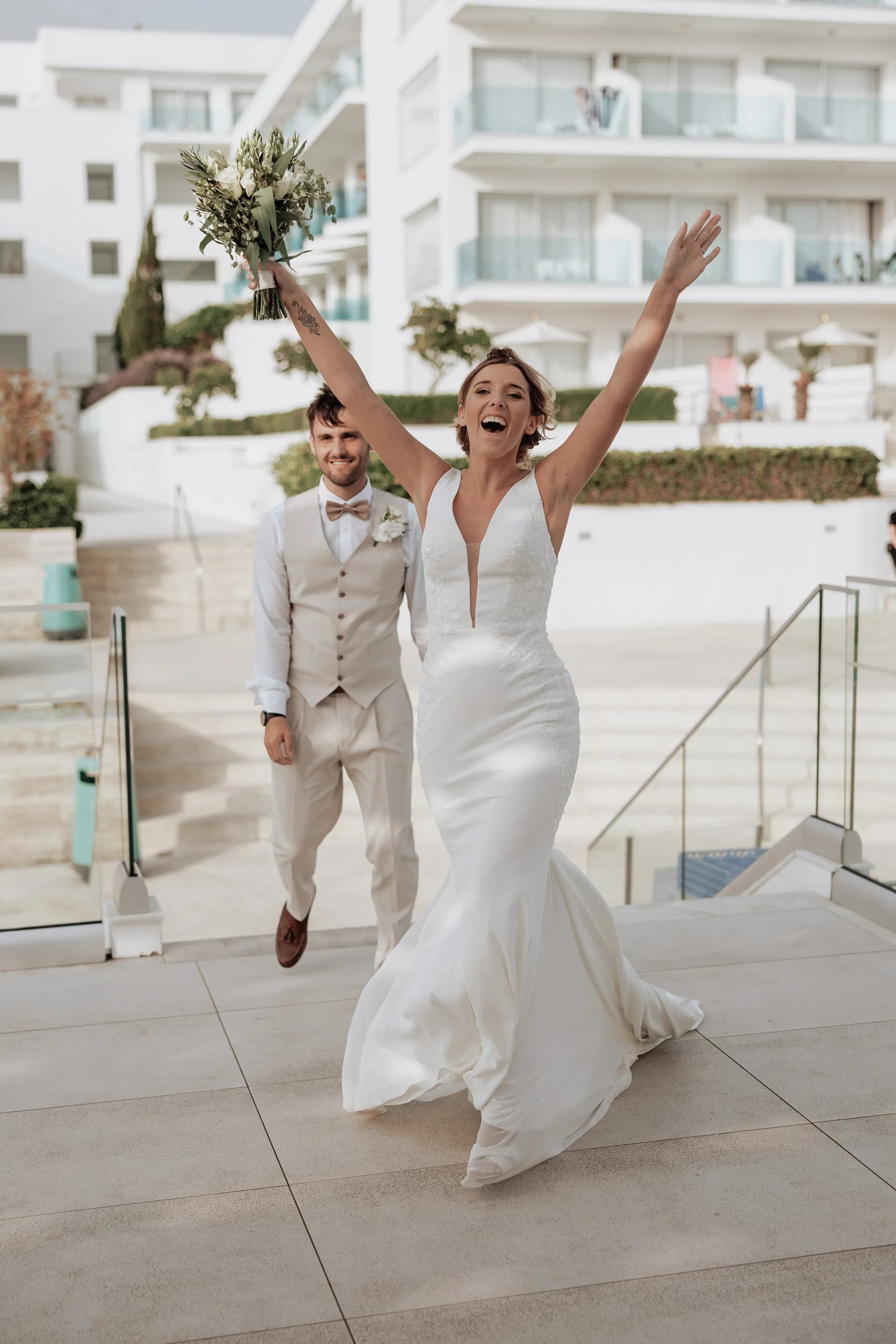 A bride entering the reception with her arms raised, King Evelthon’s white resort blocks behind