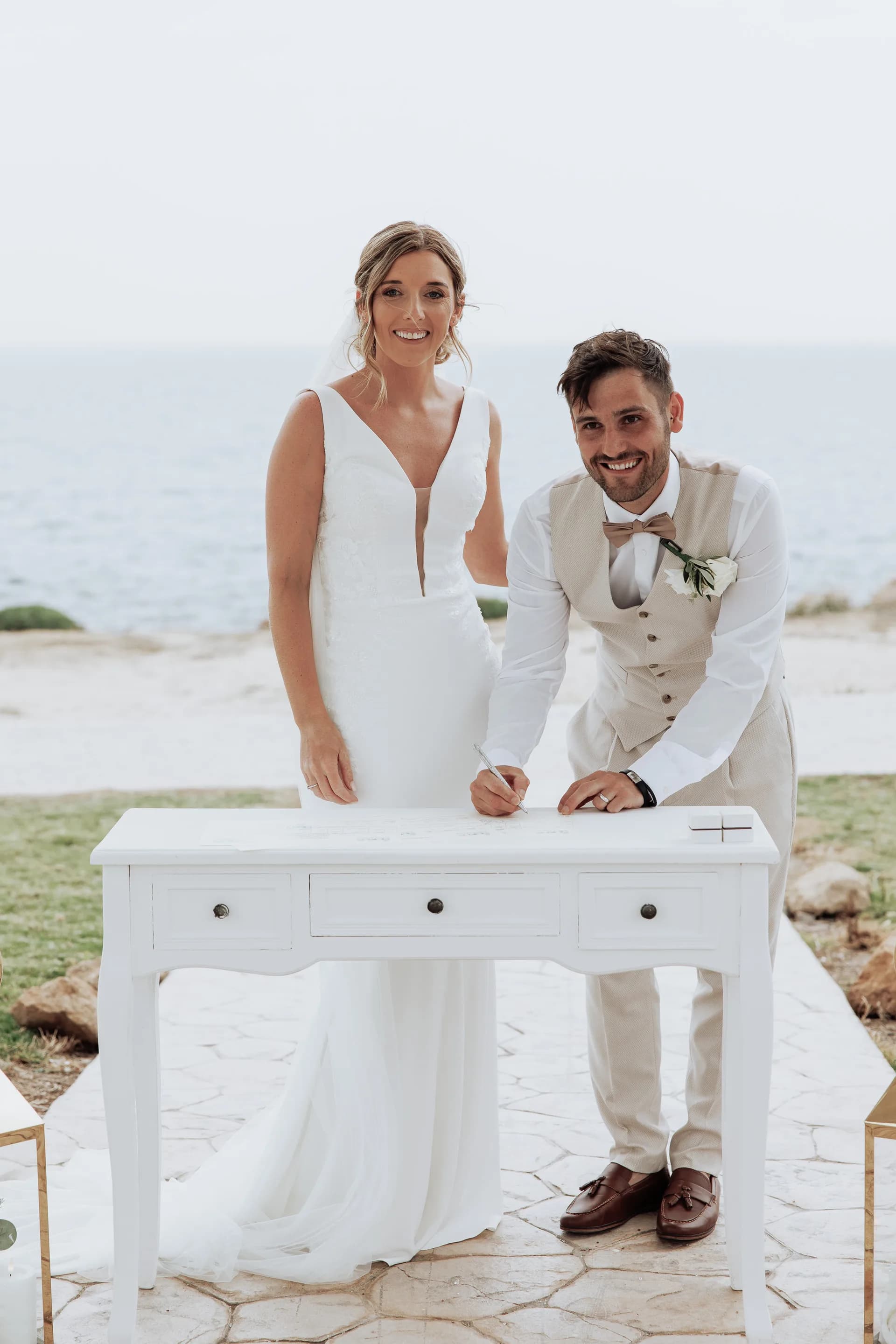 Bride and groom signing the marriage register at a white desk with the Mediterranean sea directly behind them at a Cyprus wedding