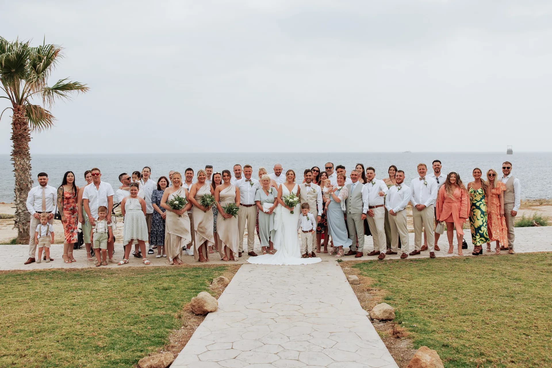 A wedding party gathered along King Evelthon’s seafront lawn beside the stone ceremony aisle