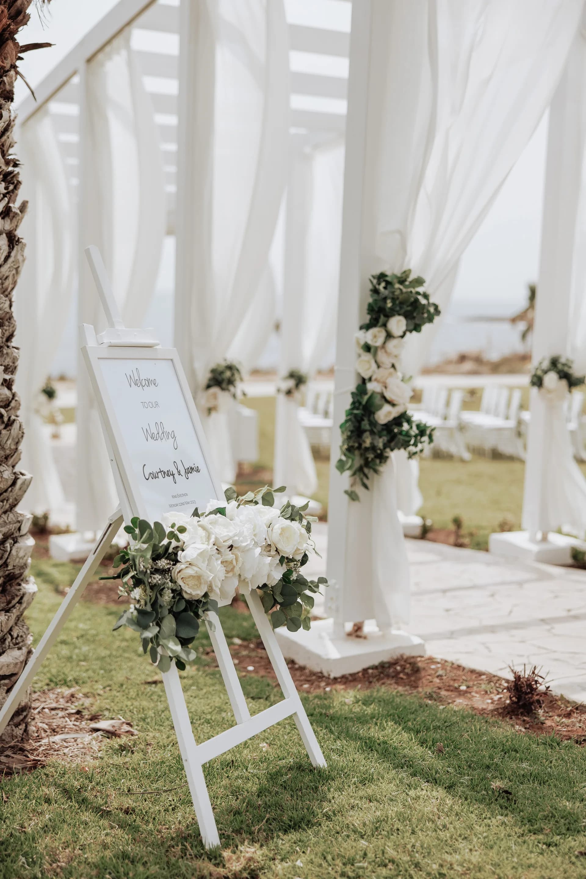 Floral-dressed welcome sign on a white easel beside King Evelthon’s ceremony drapes, lawn and palms