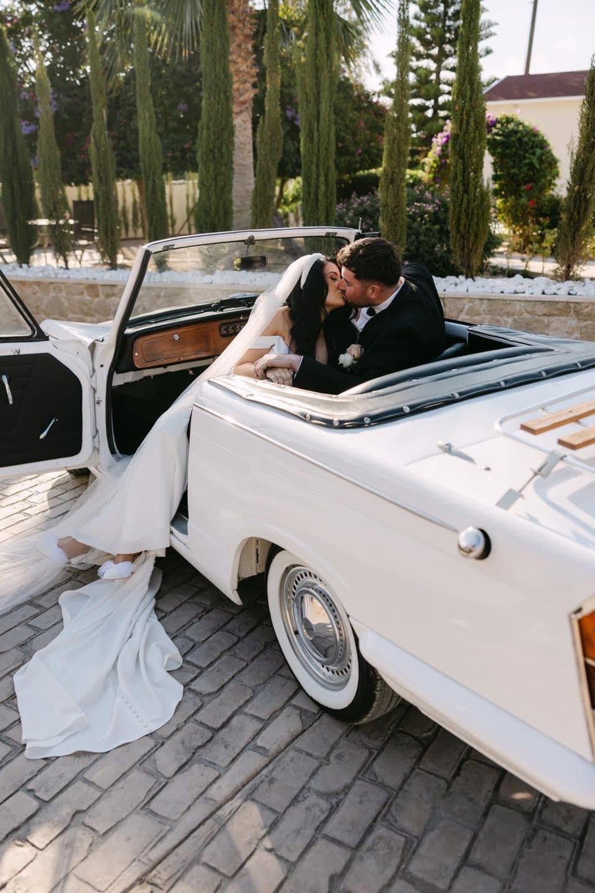 Newly married couple kissing in the back of a white vintage convertible car lined with cypress trees in Cyprus