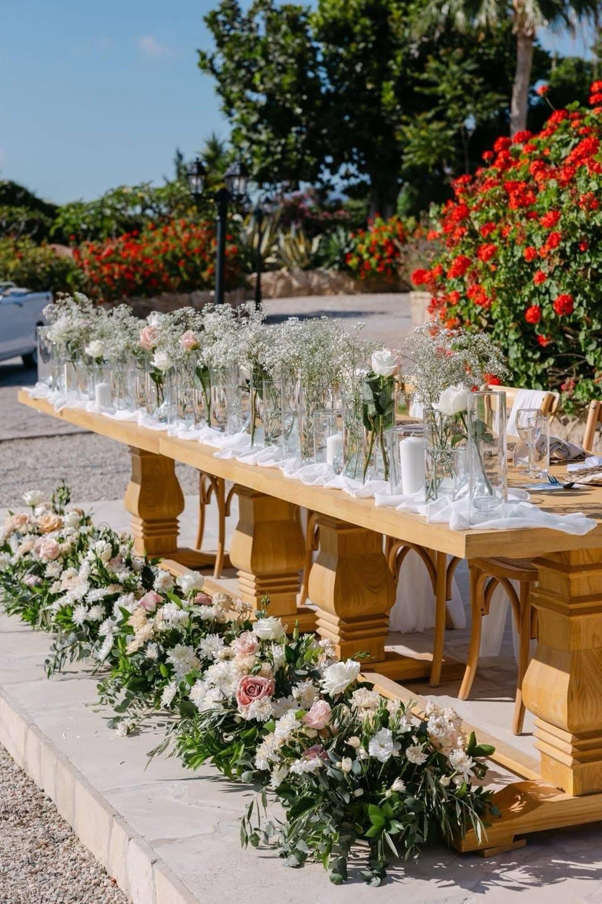 Rustic Mediterranean head table styling at L'Chateau with a long timber table, a run of glass bud vases holding gypsophila and roses, pillar candles and blush and ivory ground florals along the stone edge