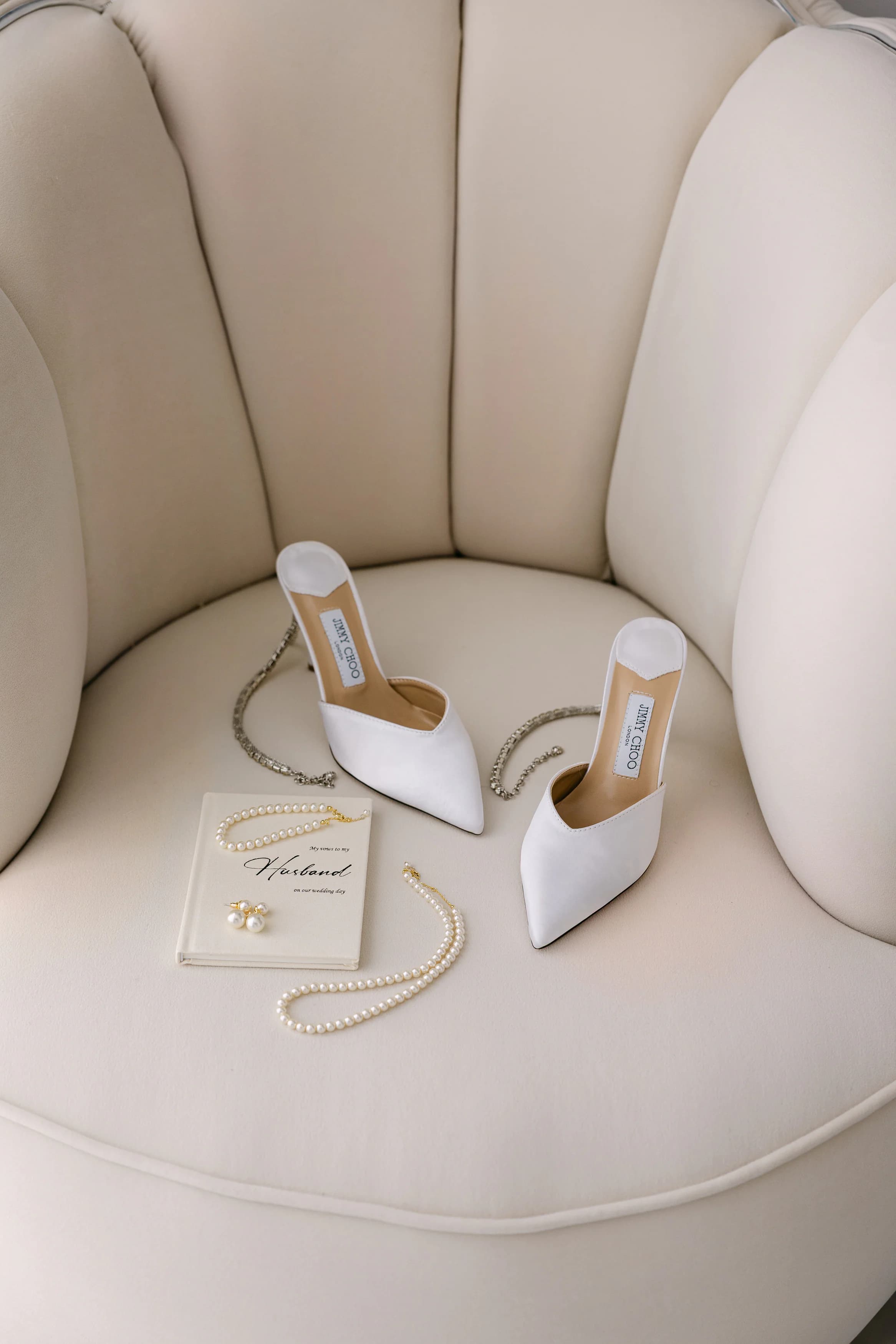 Bridal styling flatlay at OZEA — white pointed-toe heels, pearl necklace, gold earrings and personalised journal on an ivory armchair