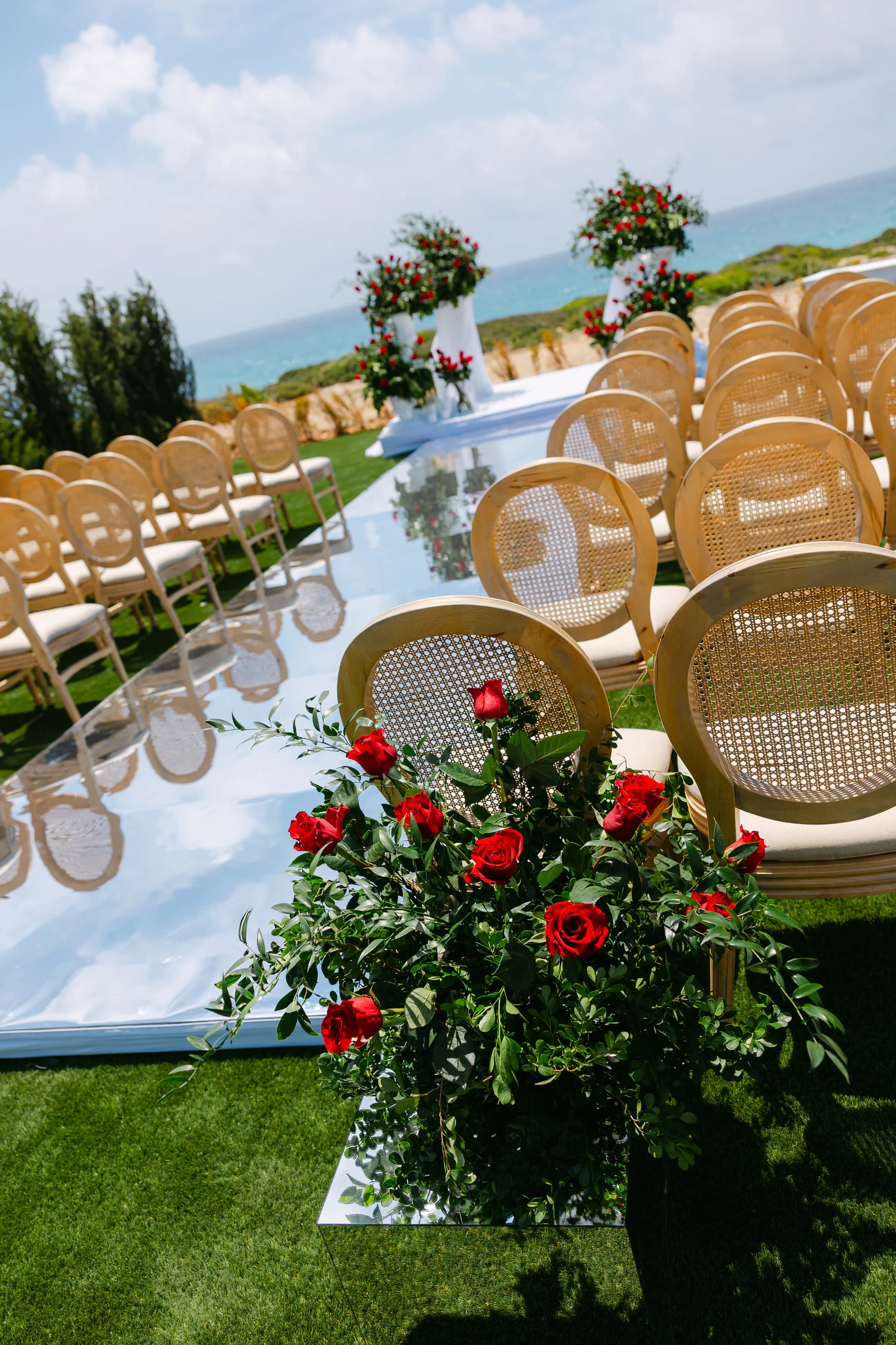Detail of the mirrored ceremony aisle at OZEA with single-stem red roses, cane chairs and reflected sky