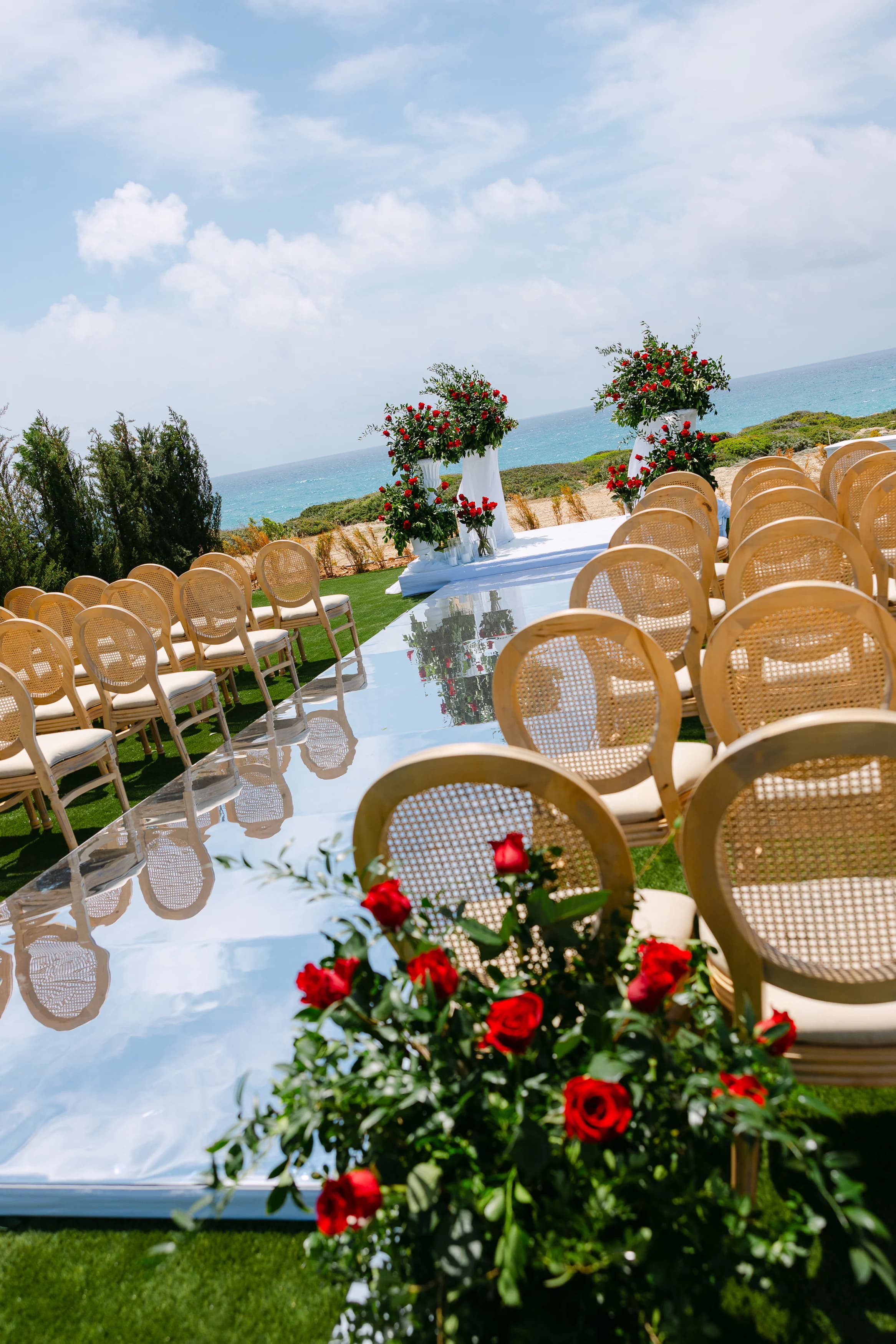 Mirrored ceremony aisle at OZEA framed by cane chairs and lush red rose arrangements with the Mediterranean Sea beyond