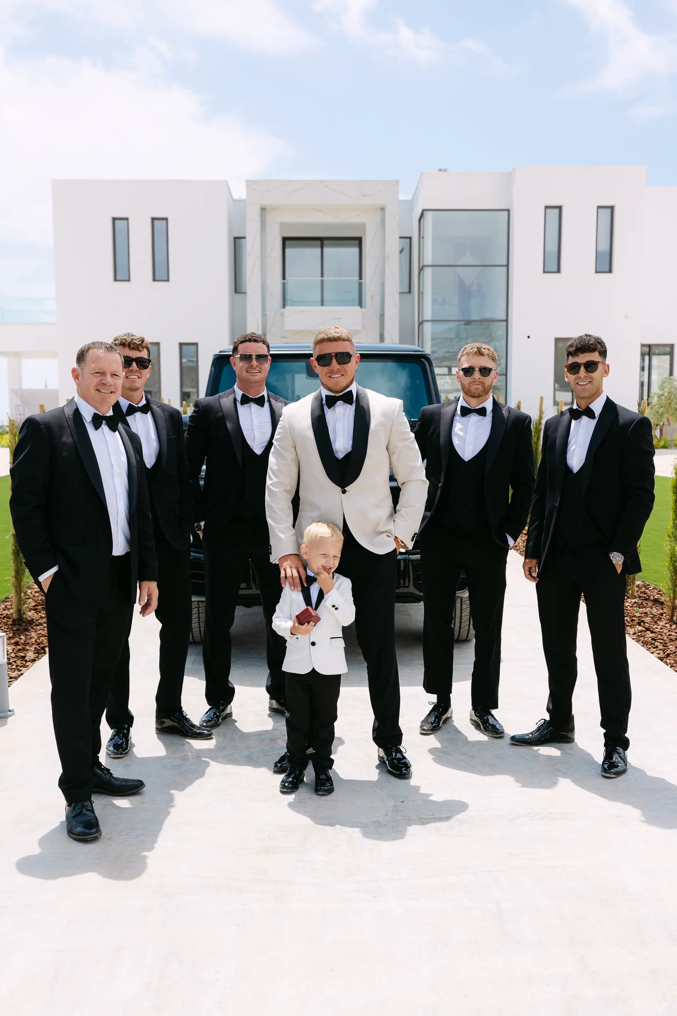 Groom Thomas and groomsmen in black tuxedos with a pageboy in front of a black Mercedes G-Wagon outside the OZEA wedding venue, Paphos
