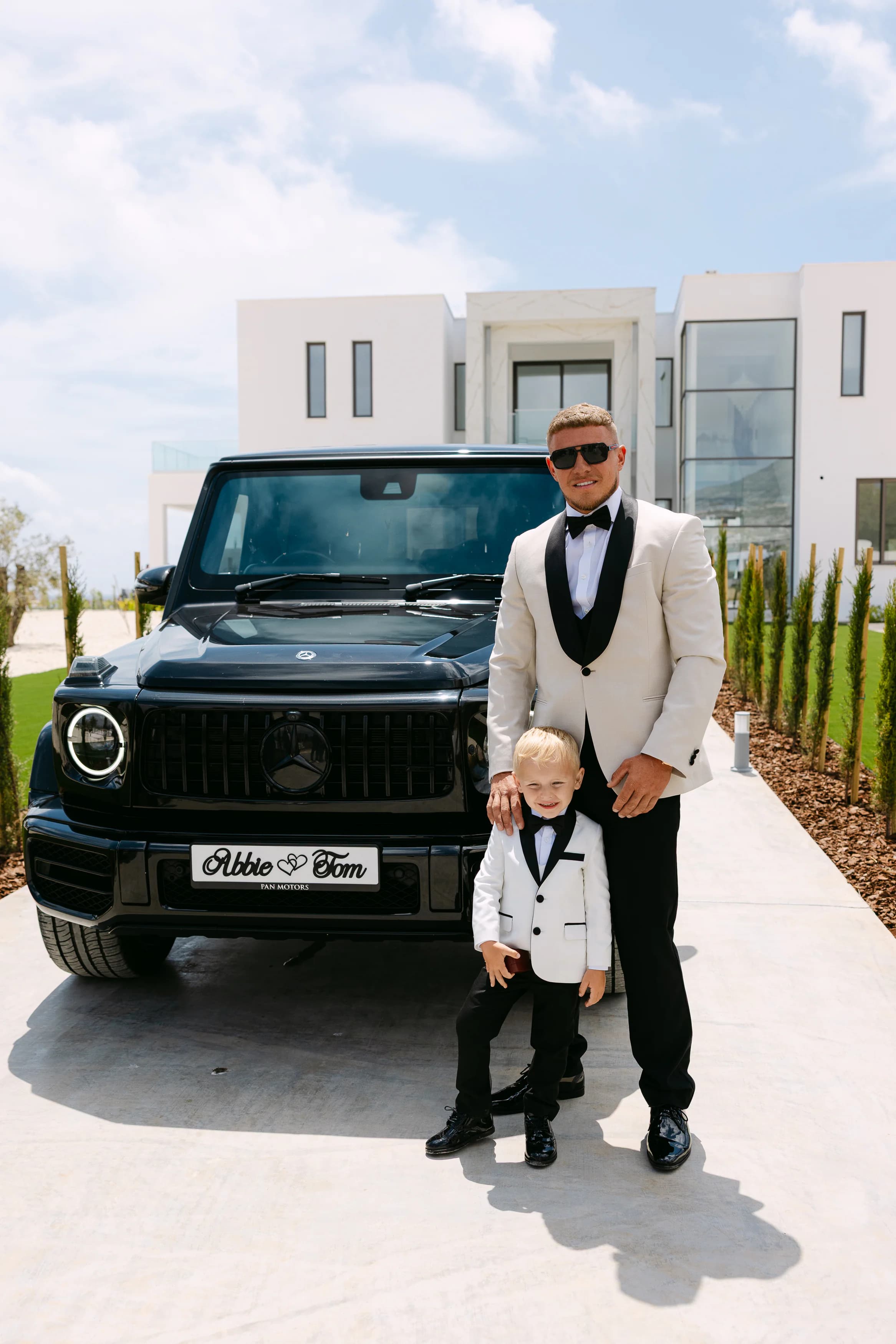 Groom Thomas in an ivory tuxedo standing beside a black Mercedes G-Wagon with a personalised Abbie & Tom number plate at OZEA