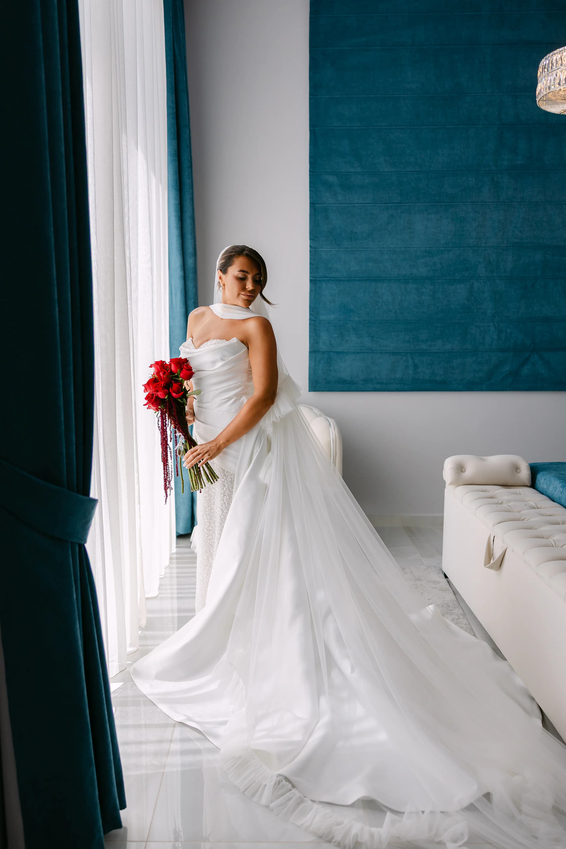 Bride in white wedding gown holding red rose bouquet by the window in the OZEA bridal suite with teal accents