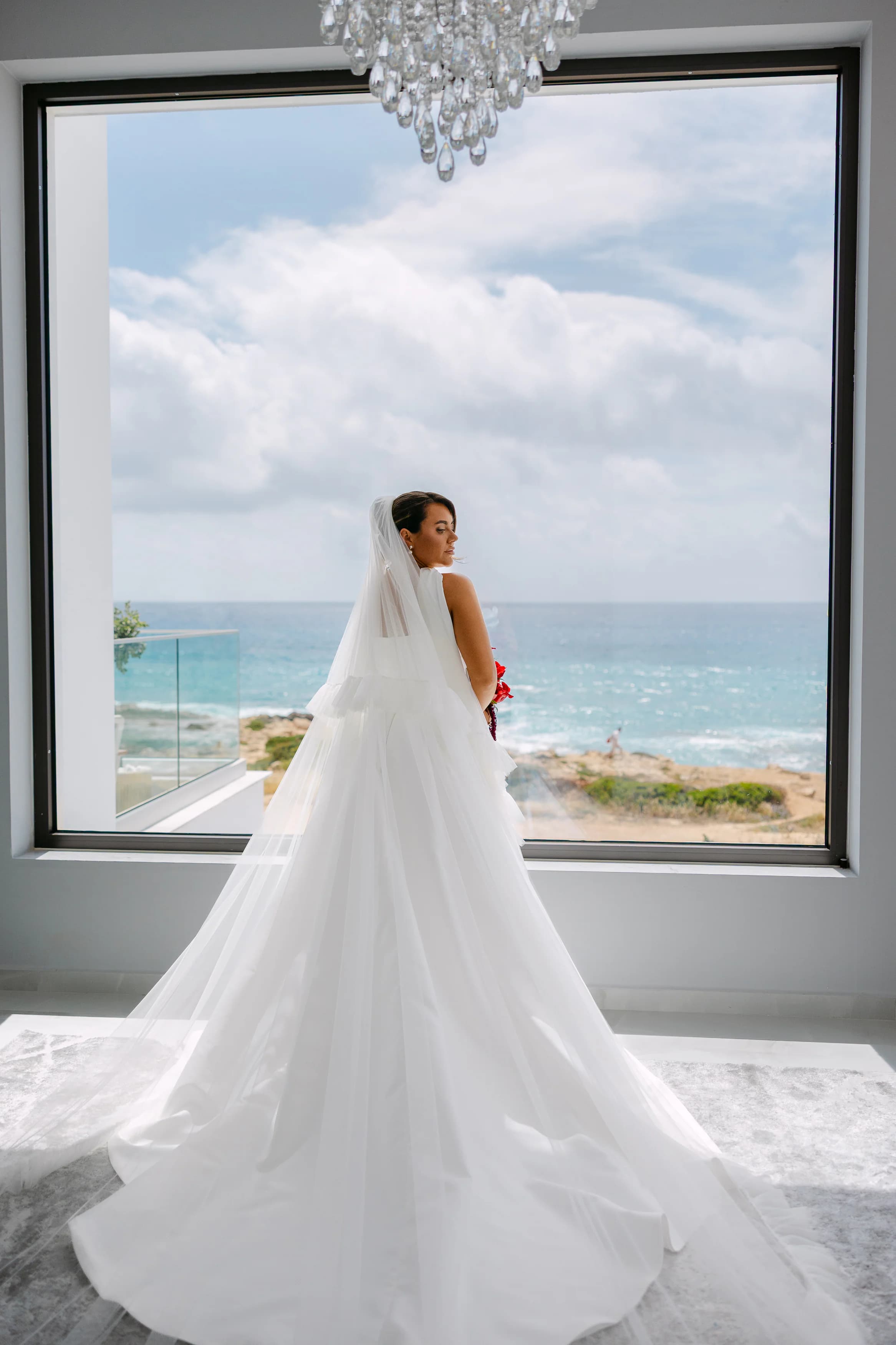 Bride in cathedral veil and ballgown looking out over the Sea Caves Paphos coastline through a full-height window at OZEA