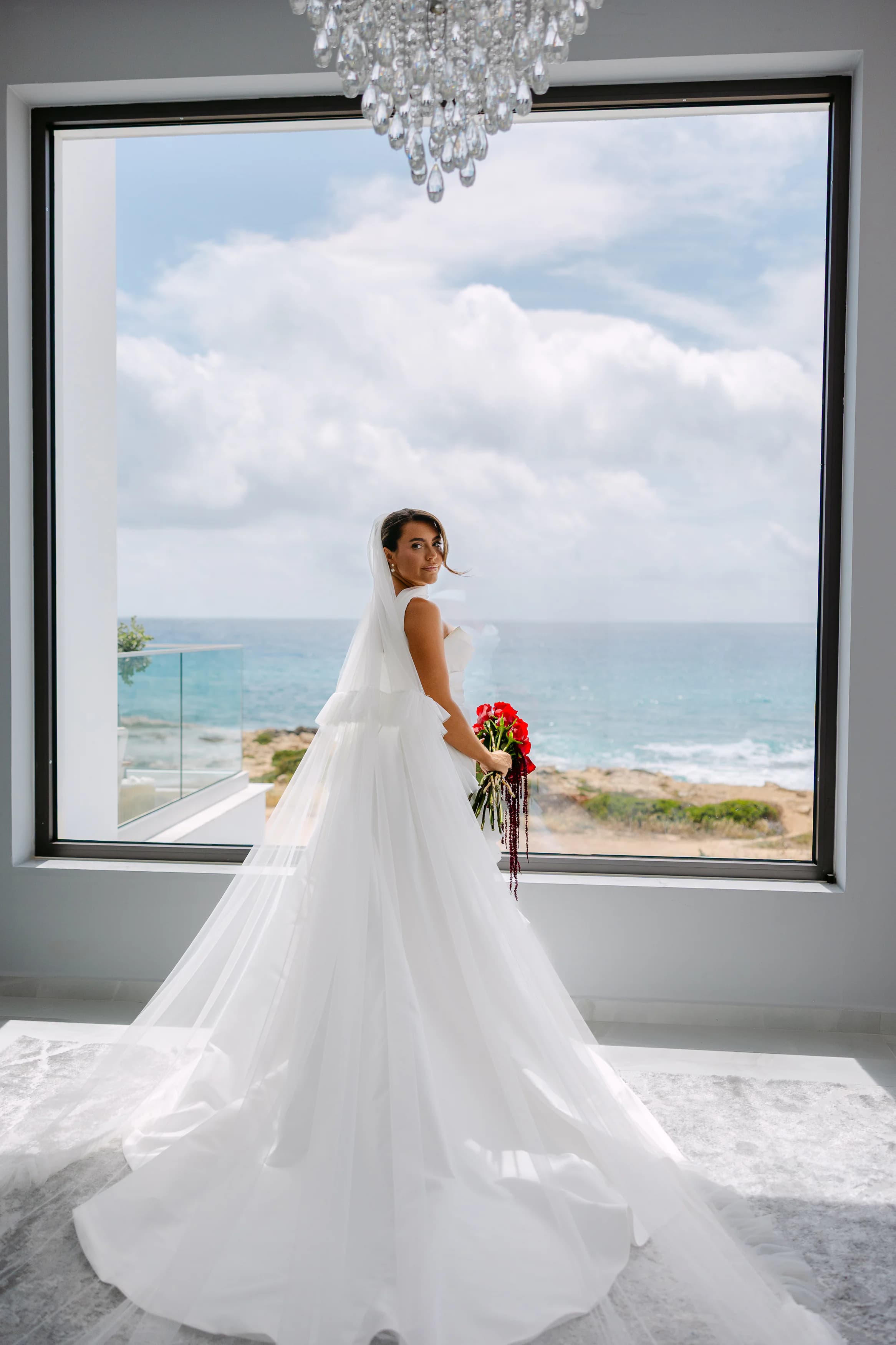 Bride in long cathedral veil from behind, framed by the OZEA sea-view window and a contemporary crystal chandelier