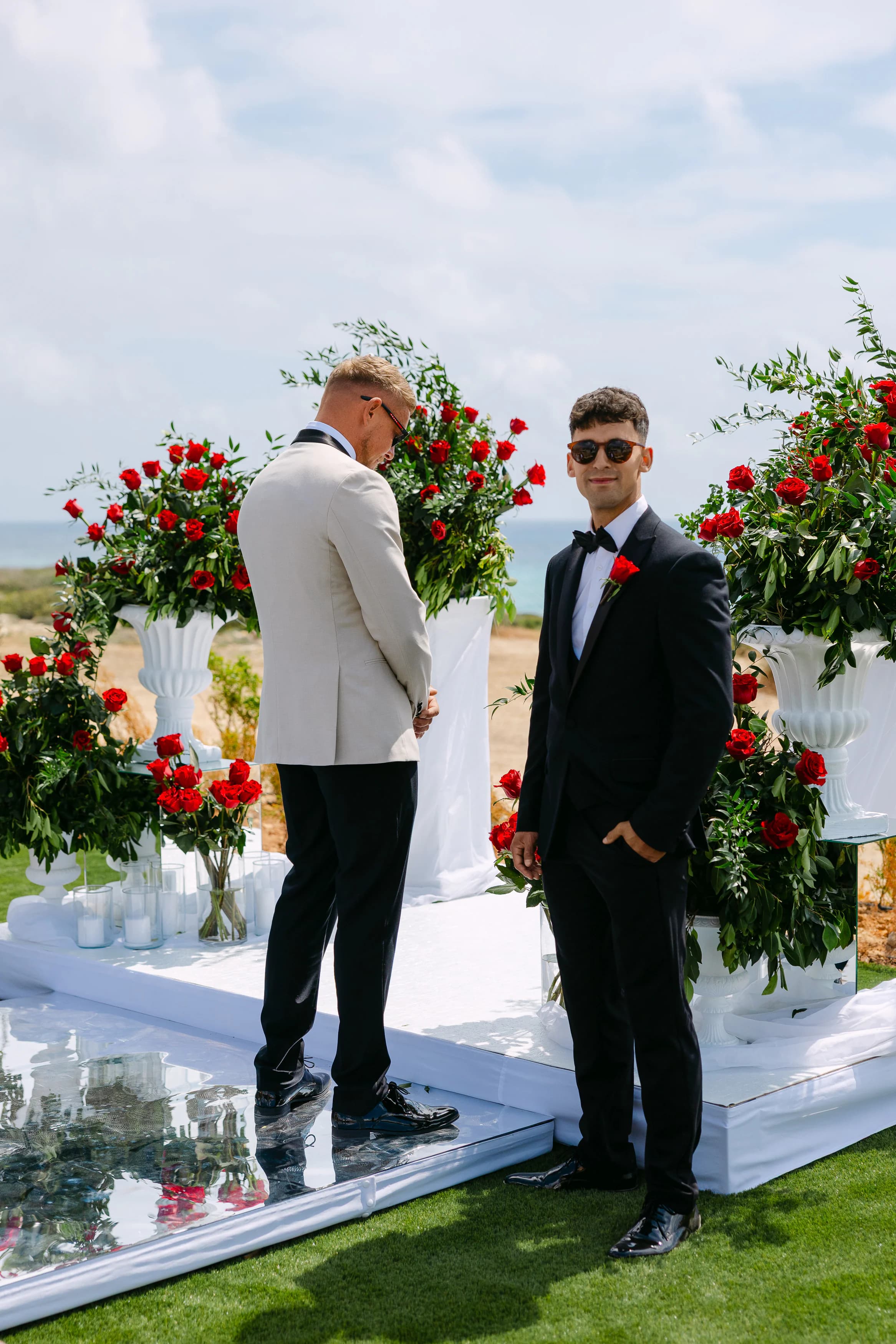 Groom and best man waiting at the OZEA ceremony altar surrounded by tall red rose arrangements with the sea behind