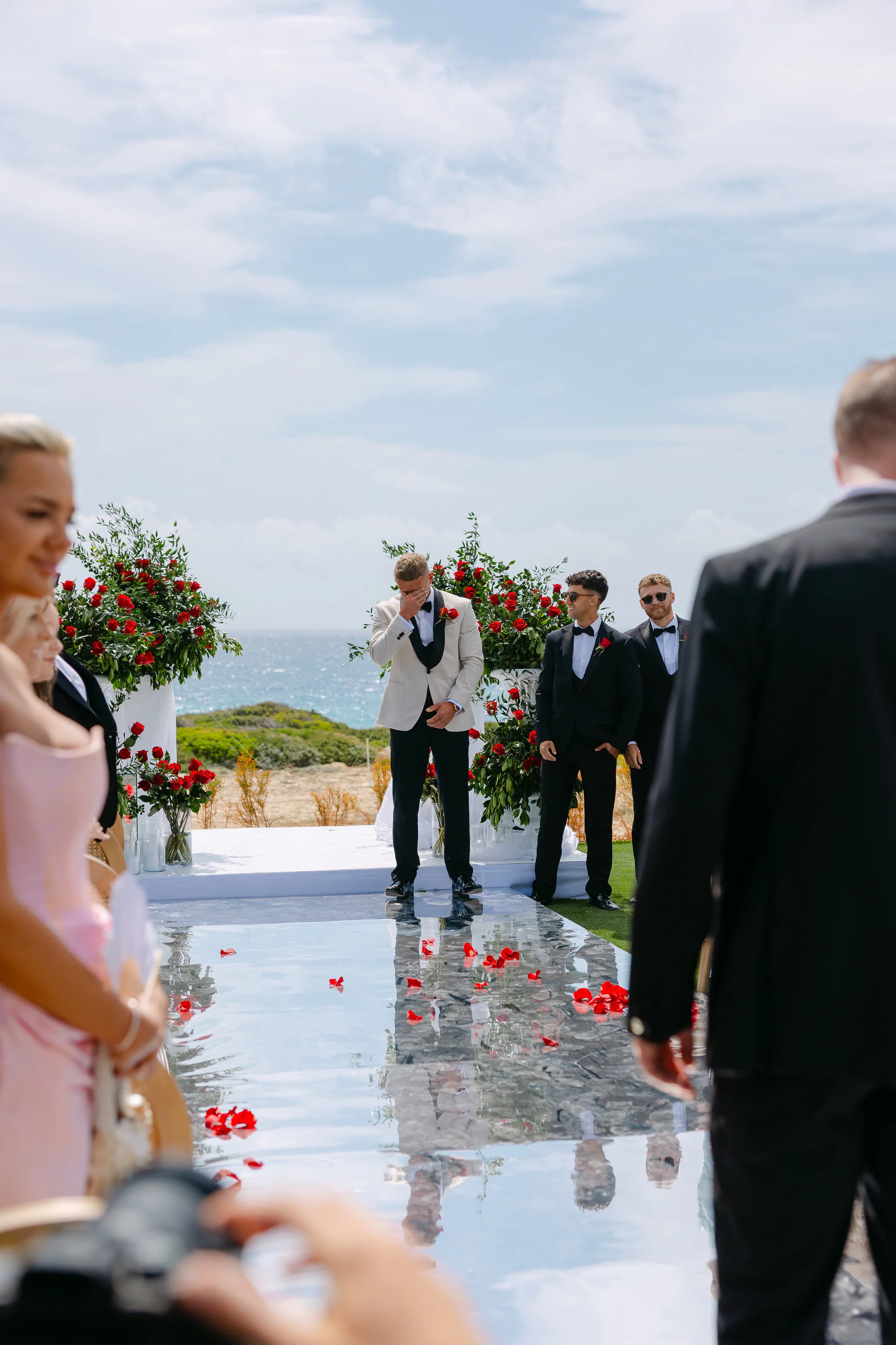 Groom Thomas wiping tears as the bride walks down the mirrored aisle at the OZEA wedding ceremony, Sea Caves Paphos