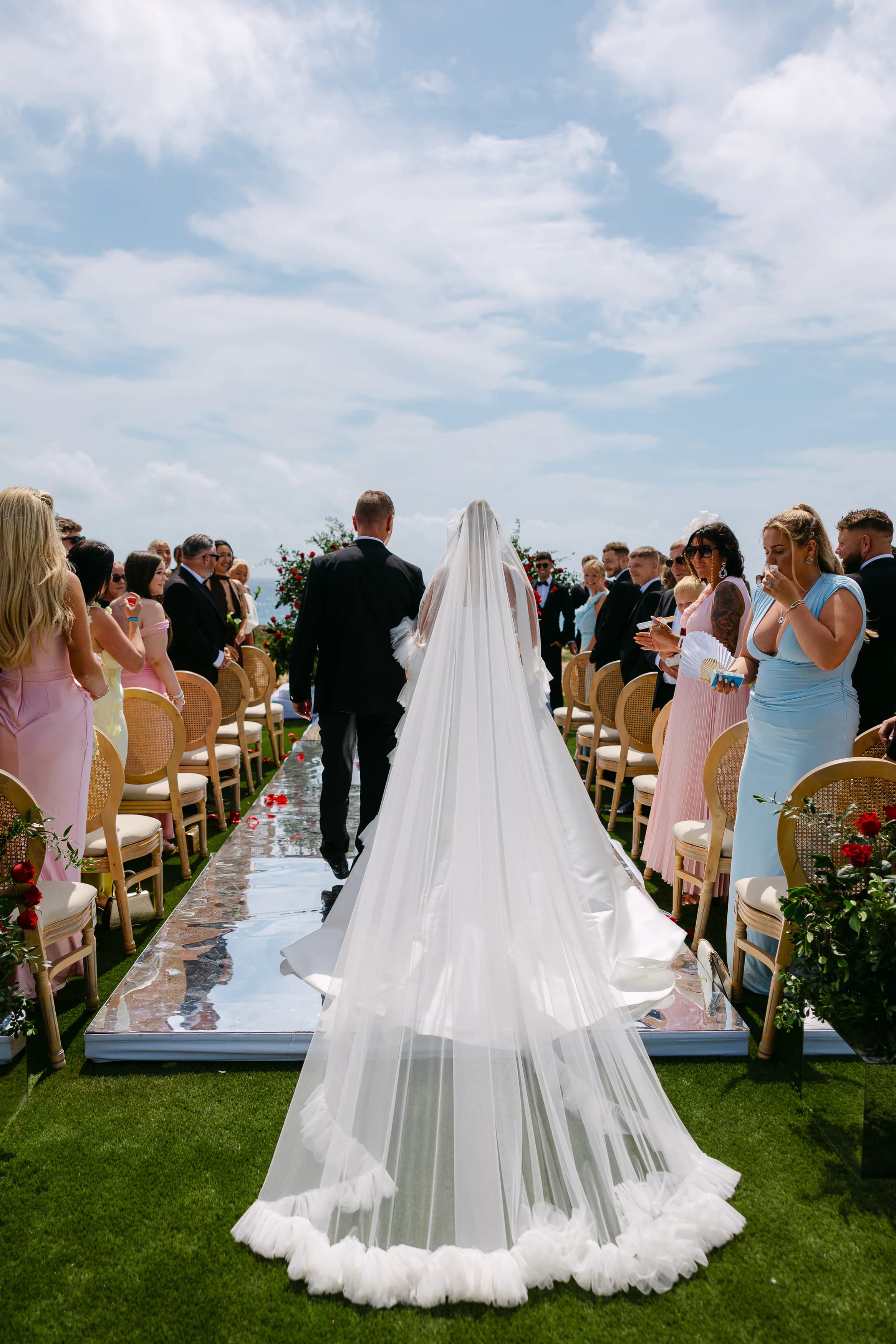 Bride walking down the OZEA wedding aisle from behind with a long cathedral veil and ruffled hem on the mirrored runner