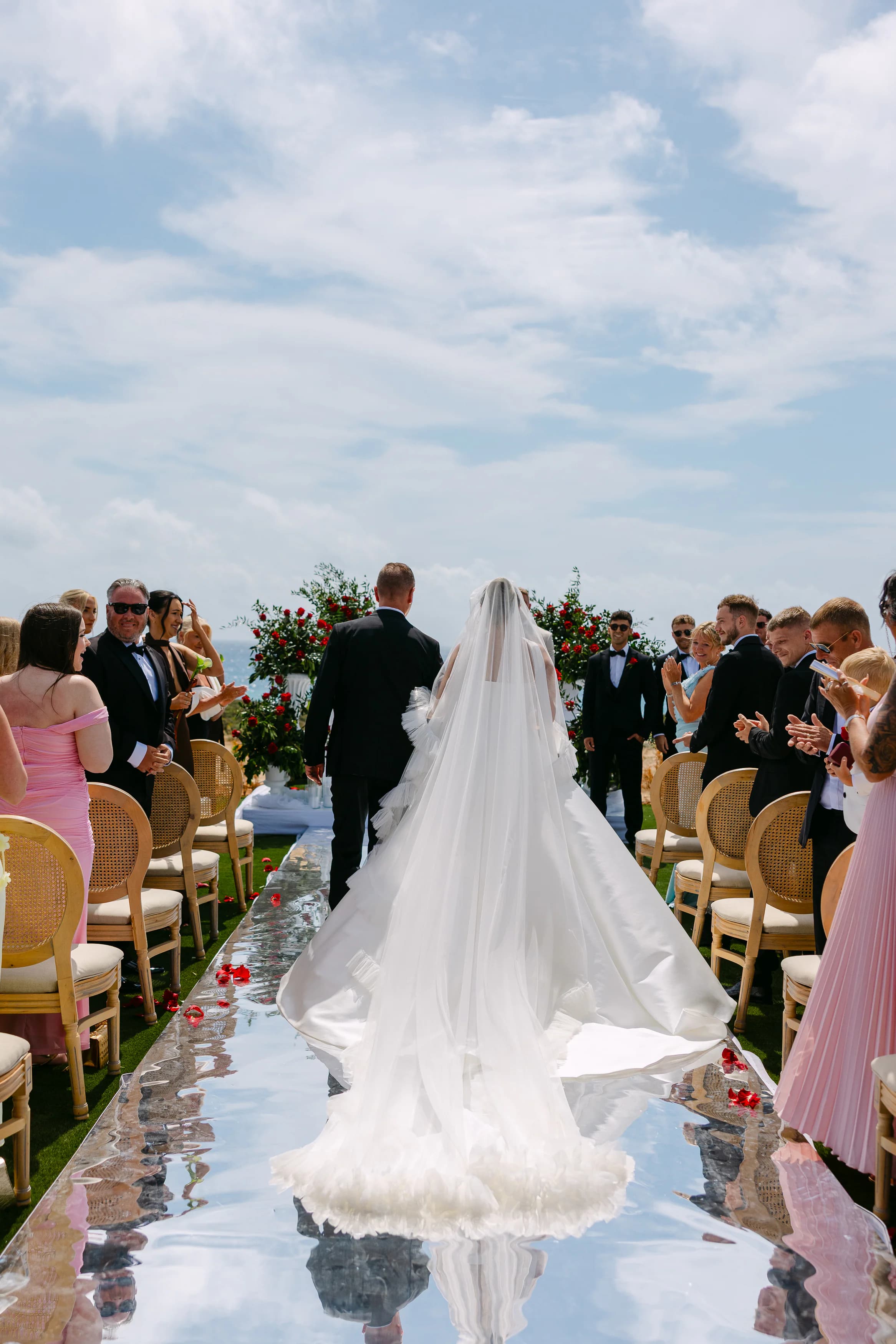 Bride walking down the mirrored aisle at OZEA with father, red rose petals scattered on the runner and guests standing