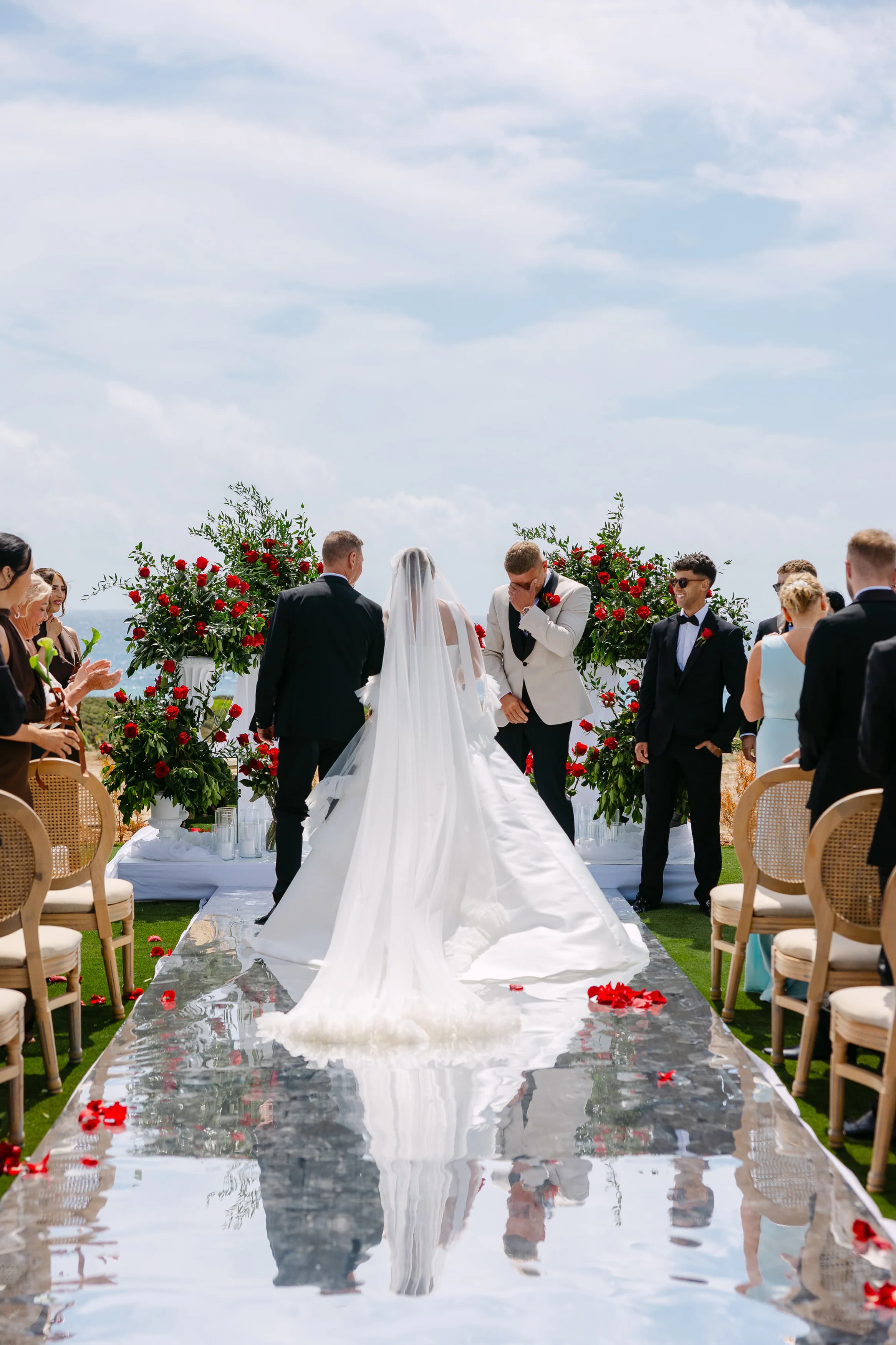 Bride walking down the mirrored ceremony aisle at OZEA Sea Caves Paphos with cathedral veil and scattered red rose petals