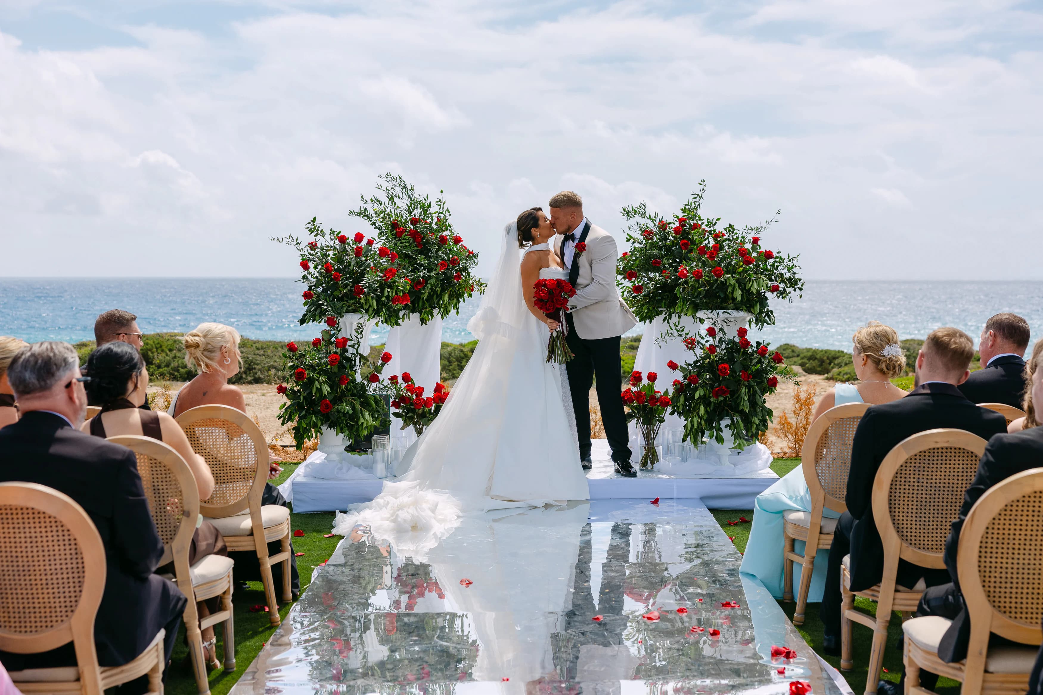 Bride and groom's first kiss at the OZEA wedding ceremony with red rose floral pedestals and Sea Caves Paphos coastline
