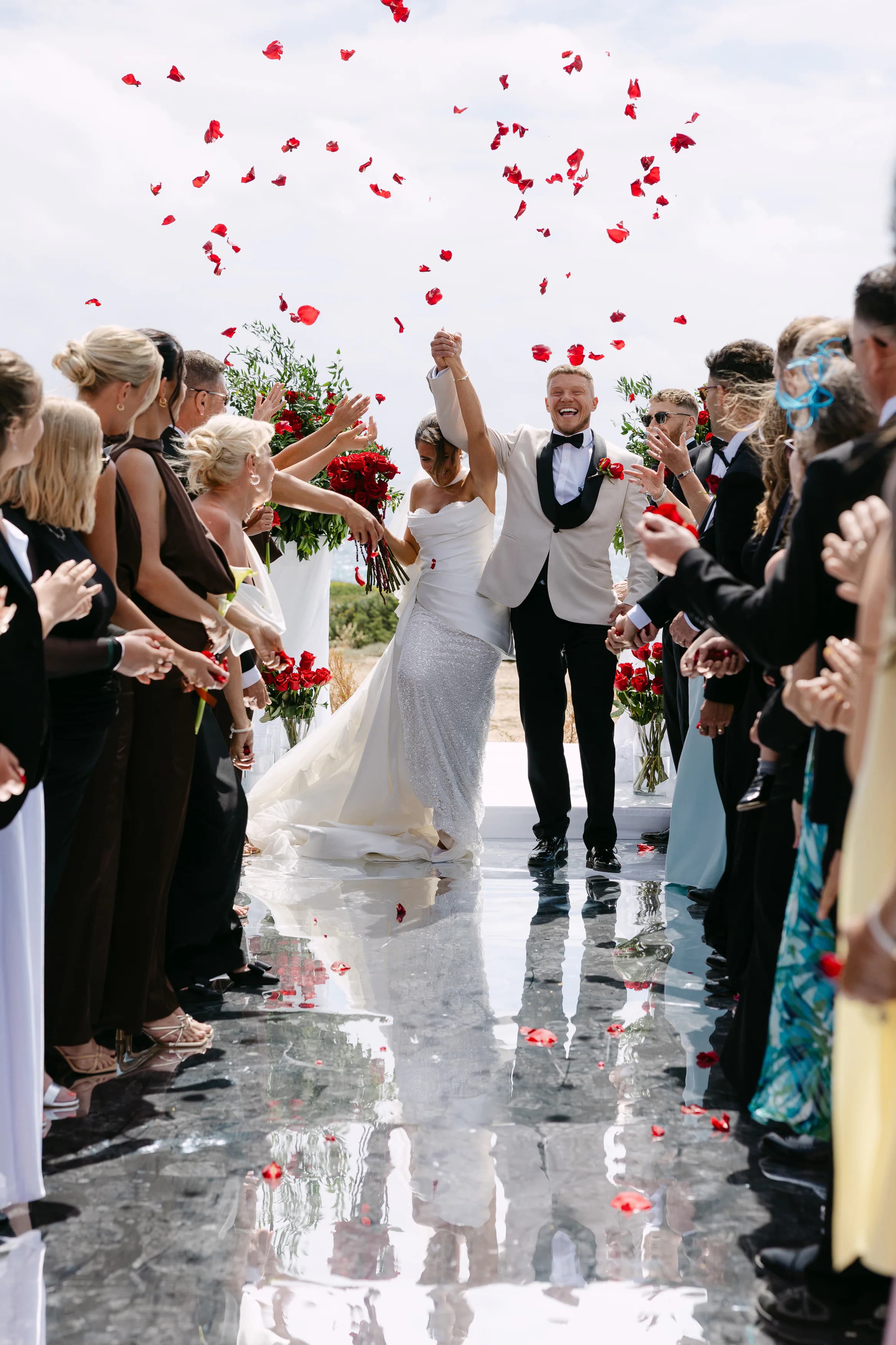 Newlyweds at OZEA celebrating with raised arms under a shower of red rose petals from guests lining the mirrored aisle