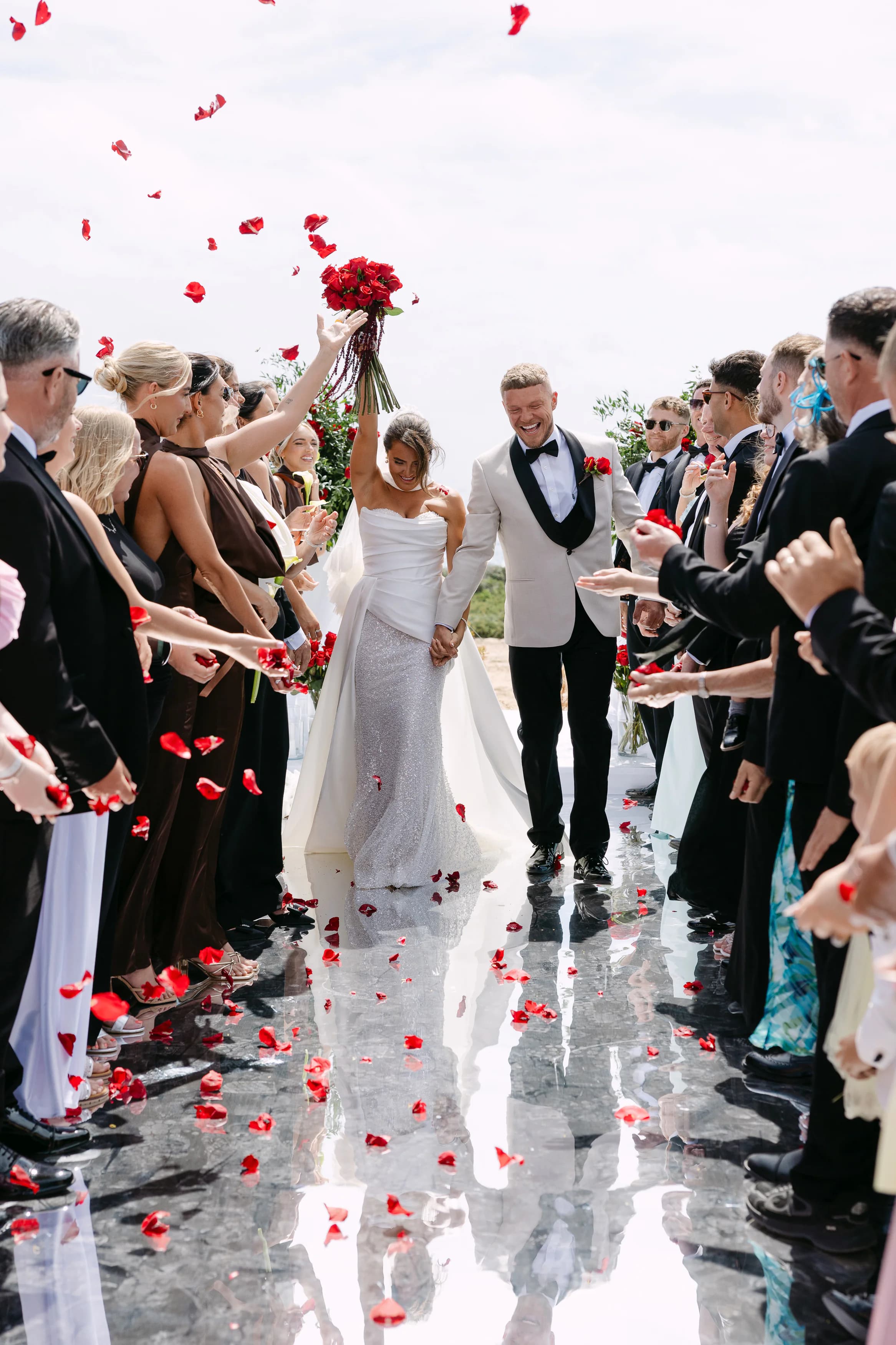 Bride and groom walking back down the mirrored aisle at OZEA with a crimson rose bouquet raised and guests showering them with petals