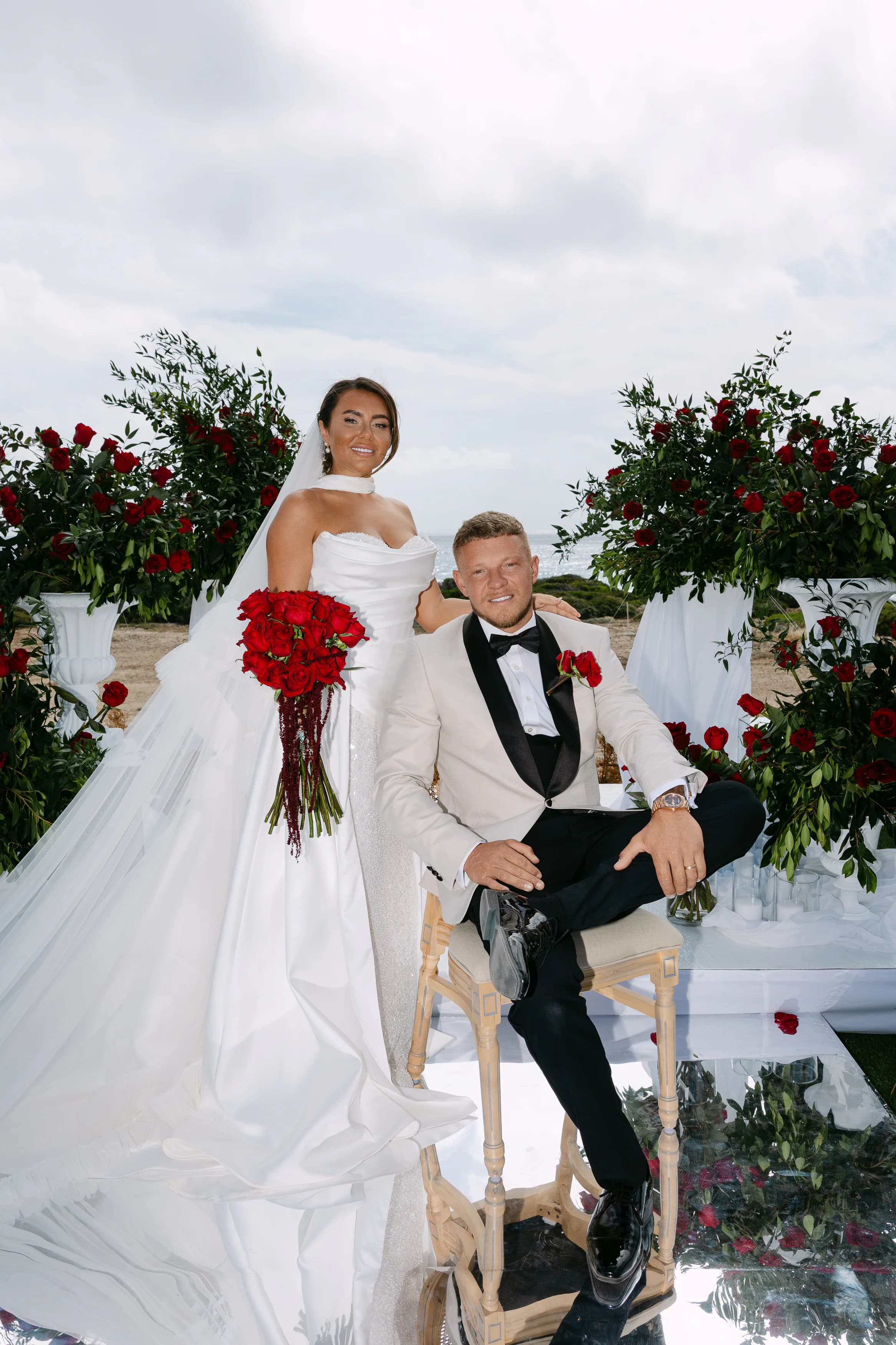 Bride and groom portrait at the OZEA ceremony altar framed by tall red rose pedestals — classic black tuxedo and ivory ballgown
