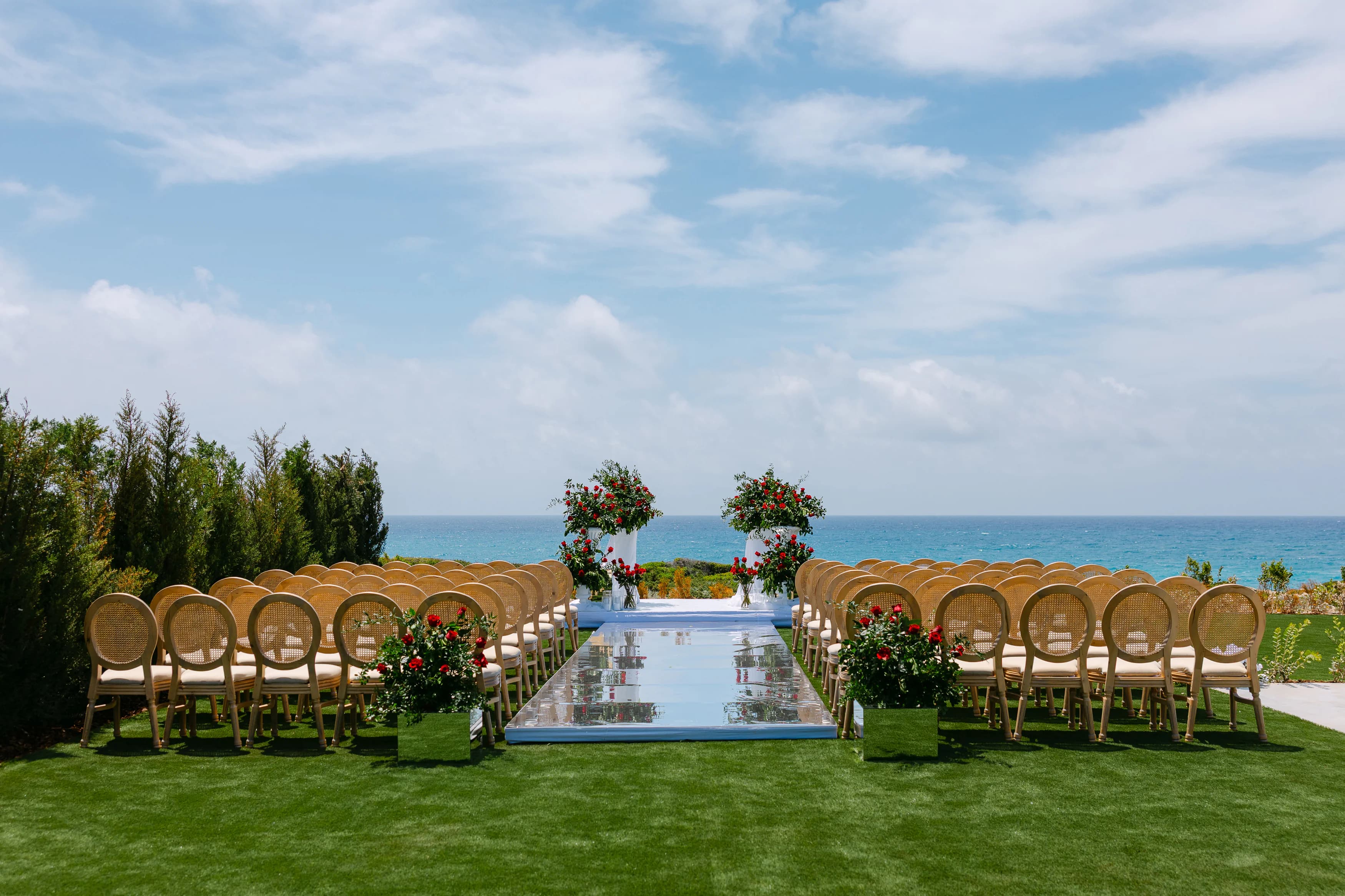 OZEA outdoor wedding ceremony setup at Sea Caves Paphos — mirrored aisle runner, cane chairs, red rose altar pedestals and open Mediterranean sea views