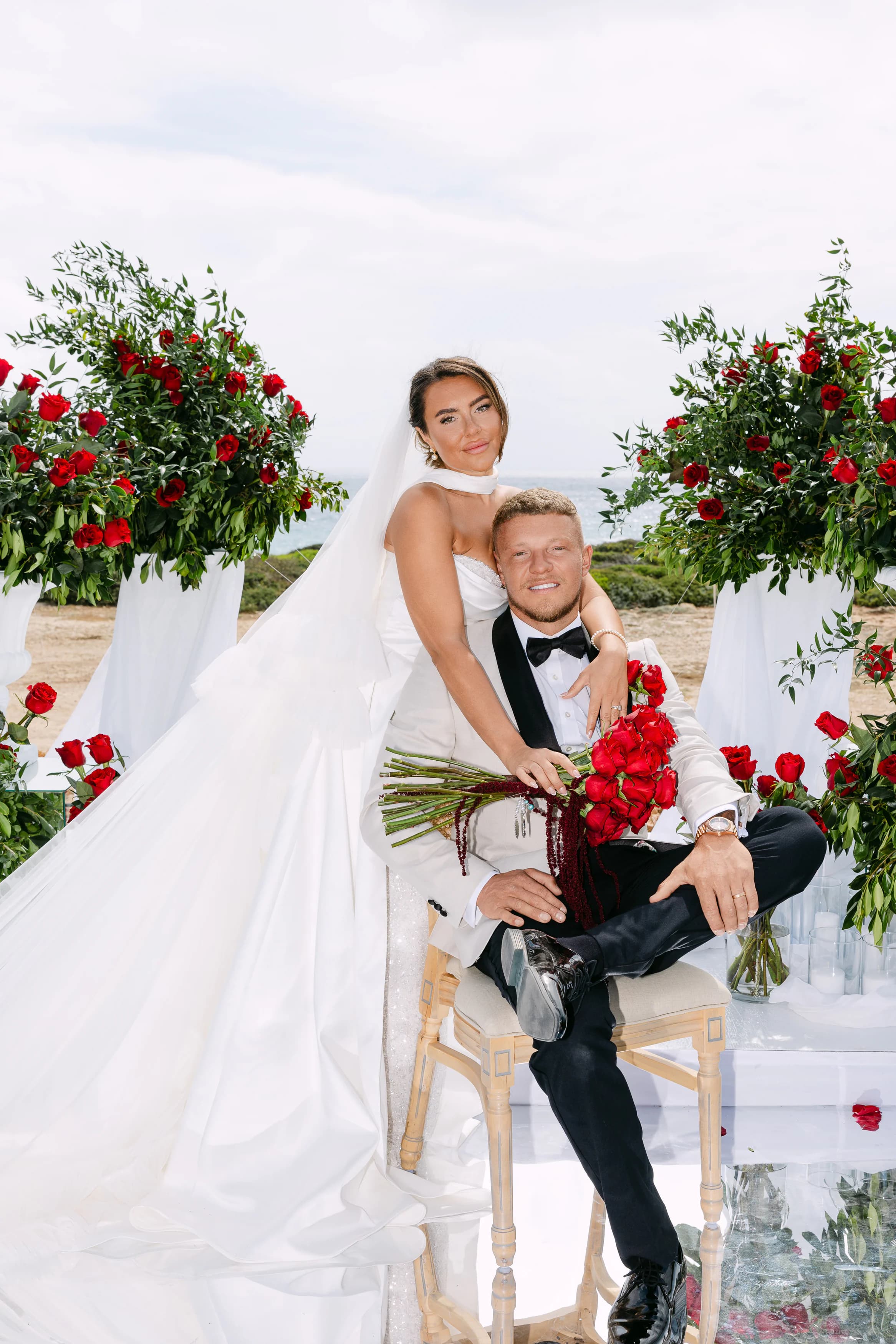 Bride leaning over seated groom with a crimson rose bouquet between them at the OZEA ceremony space, Sea Caves Paphos
