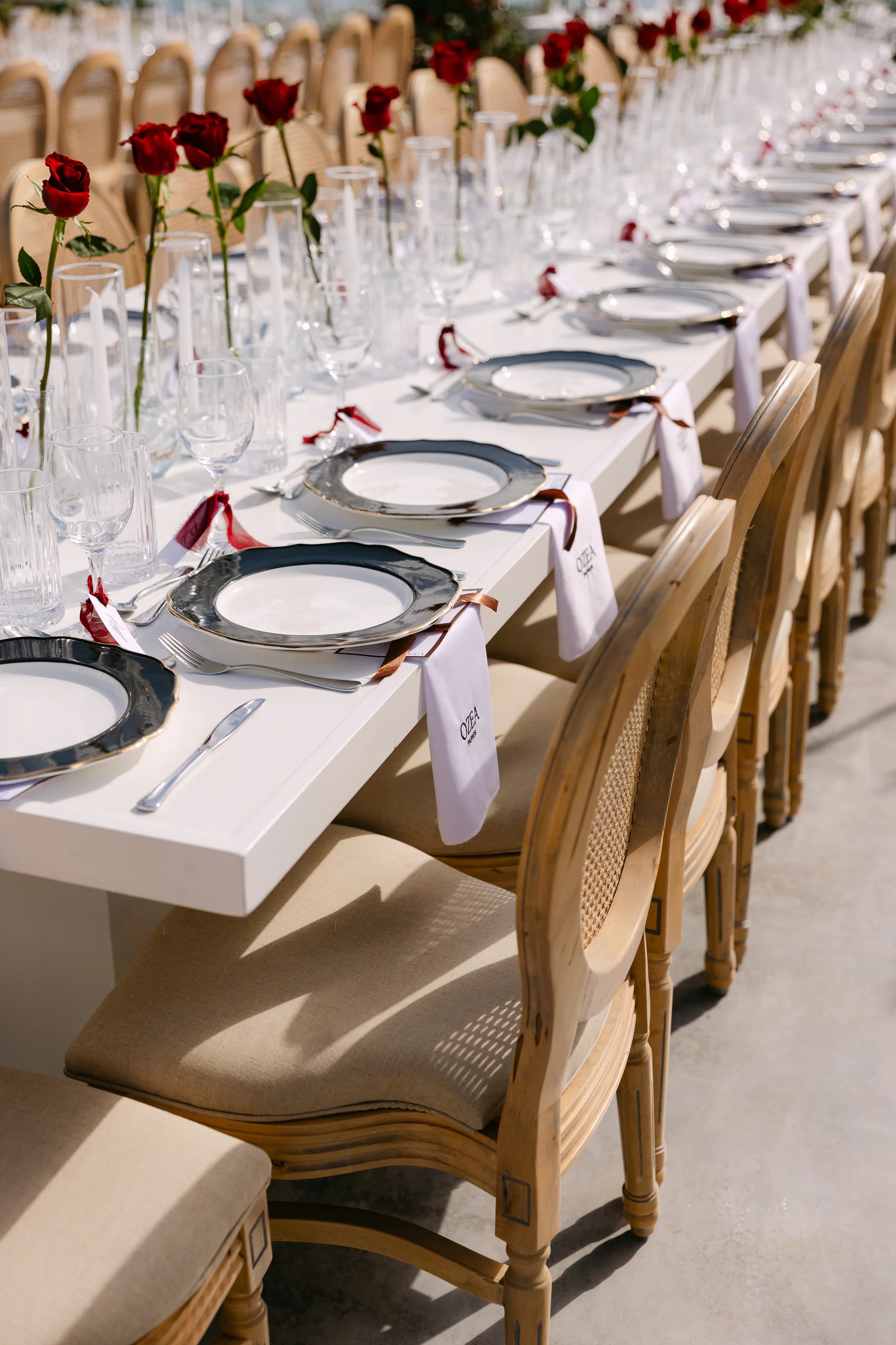 Detail of the OZEA wedding reception table — silver-rimmed charger plates, ribbon-tied napkins with monogrammed name cards and single red roses in glass flutes