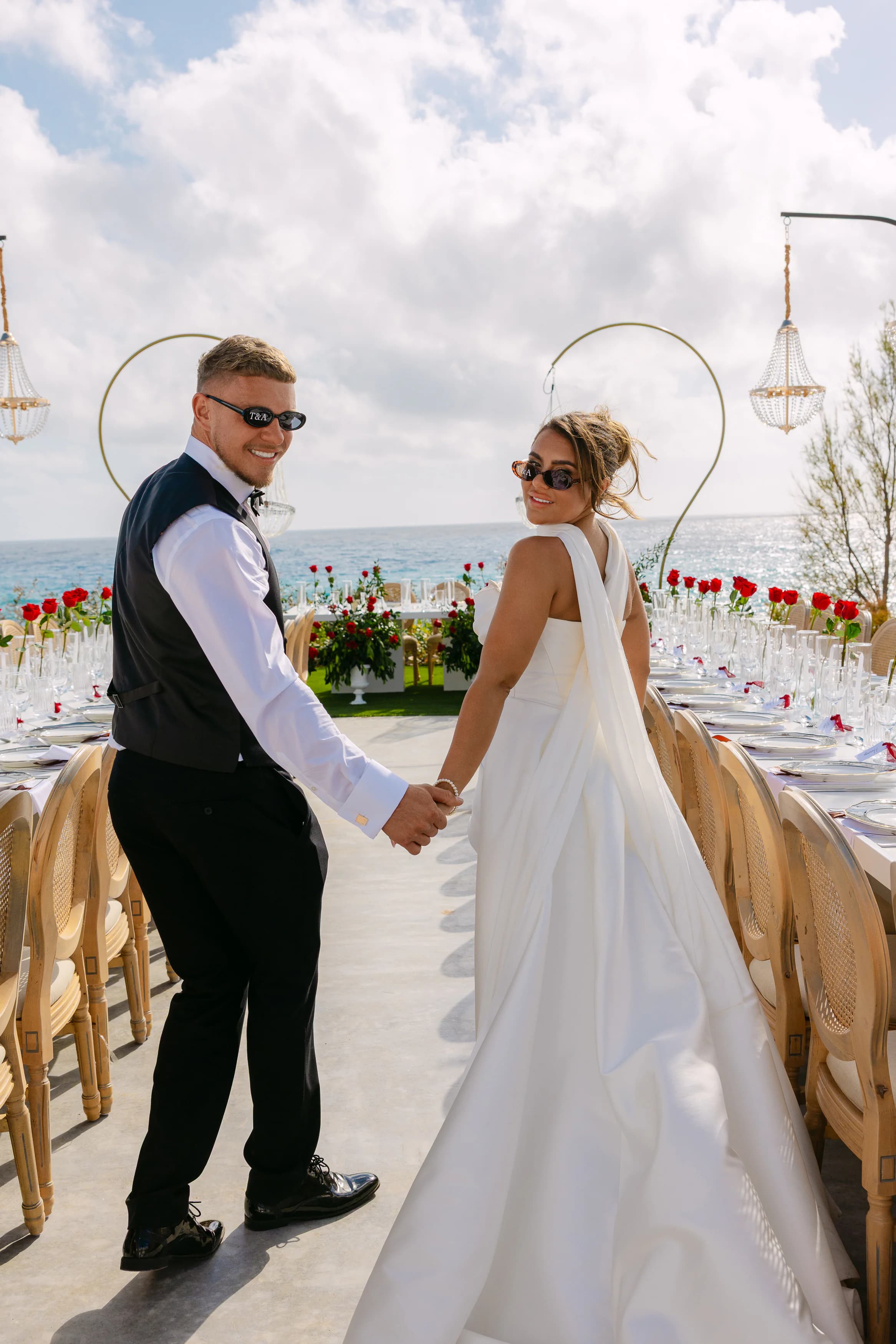 Newlyweds Abbie and Thomas walking hand in hand toward the OZEA reception table with crystal chandeliers and the sea behind