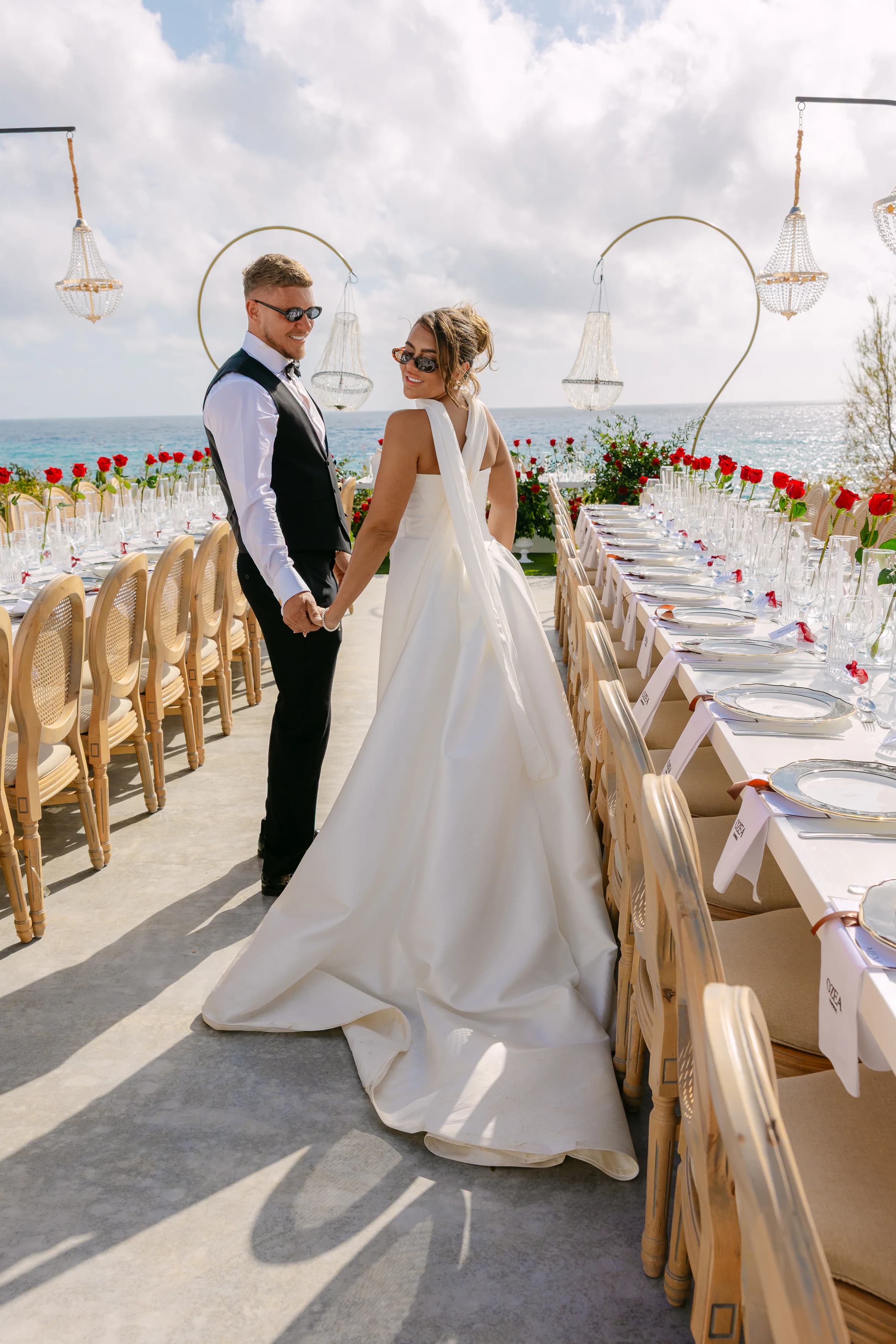 Bride and groom holding hands beside the OZEA wedding banquet table with red rose runner and chandeliers overhead