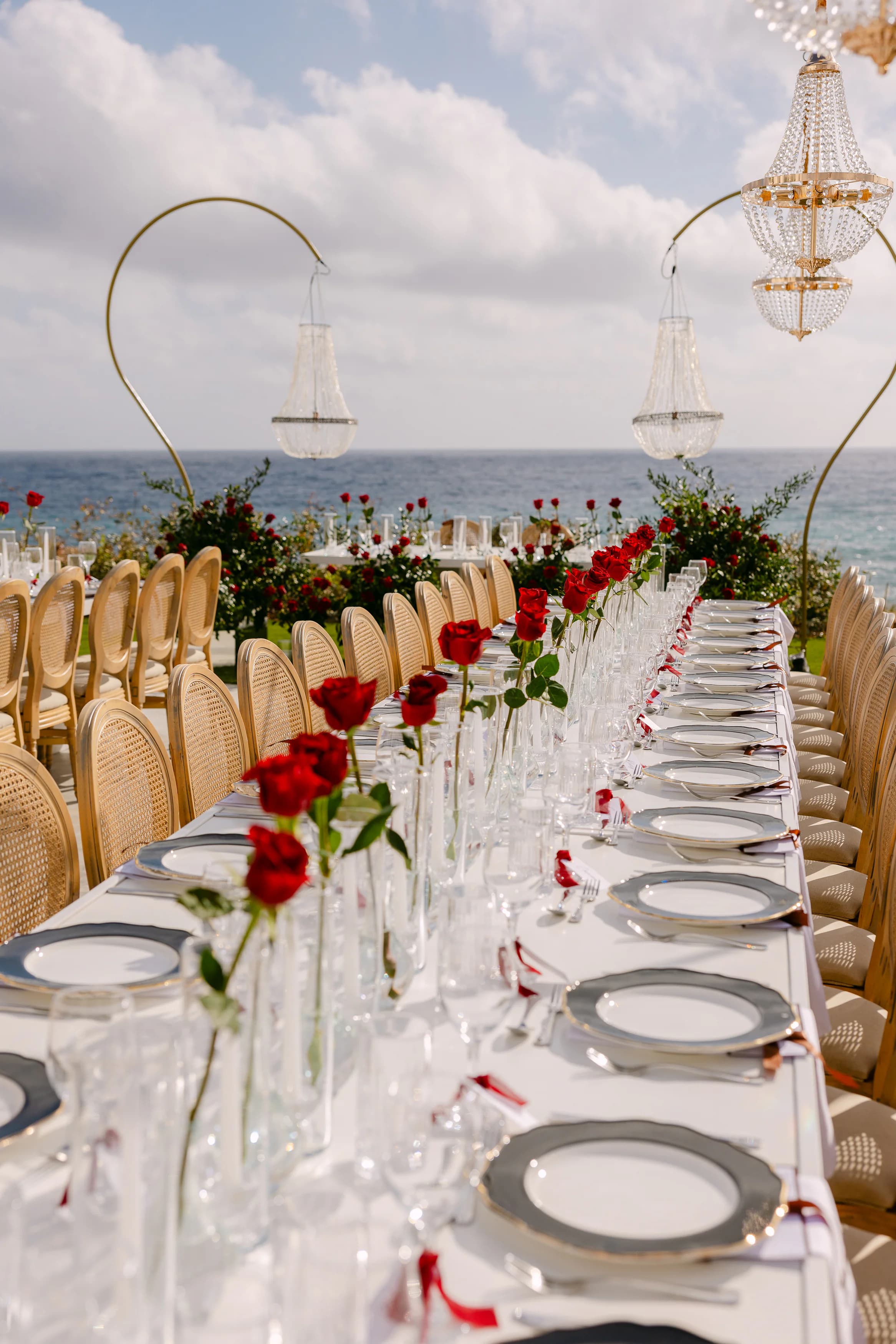 OZEA wedding banquet table styled with single red roses in flutes, crystal stemware and silver-rimmed place settings against the sea