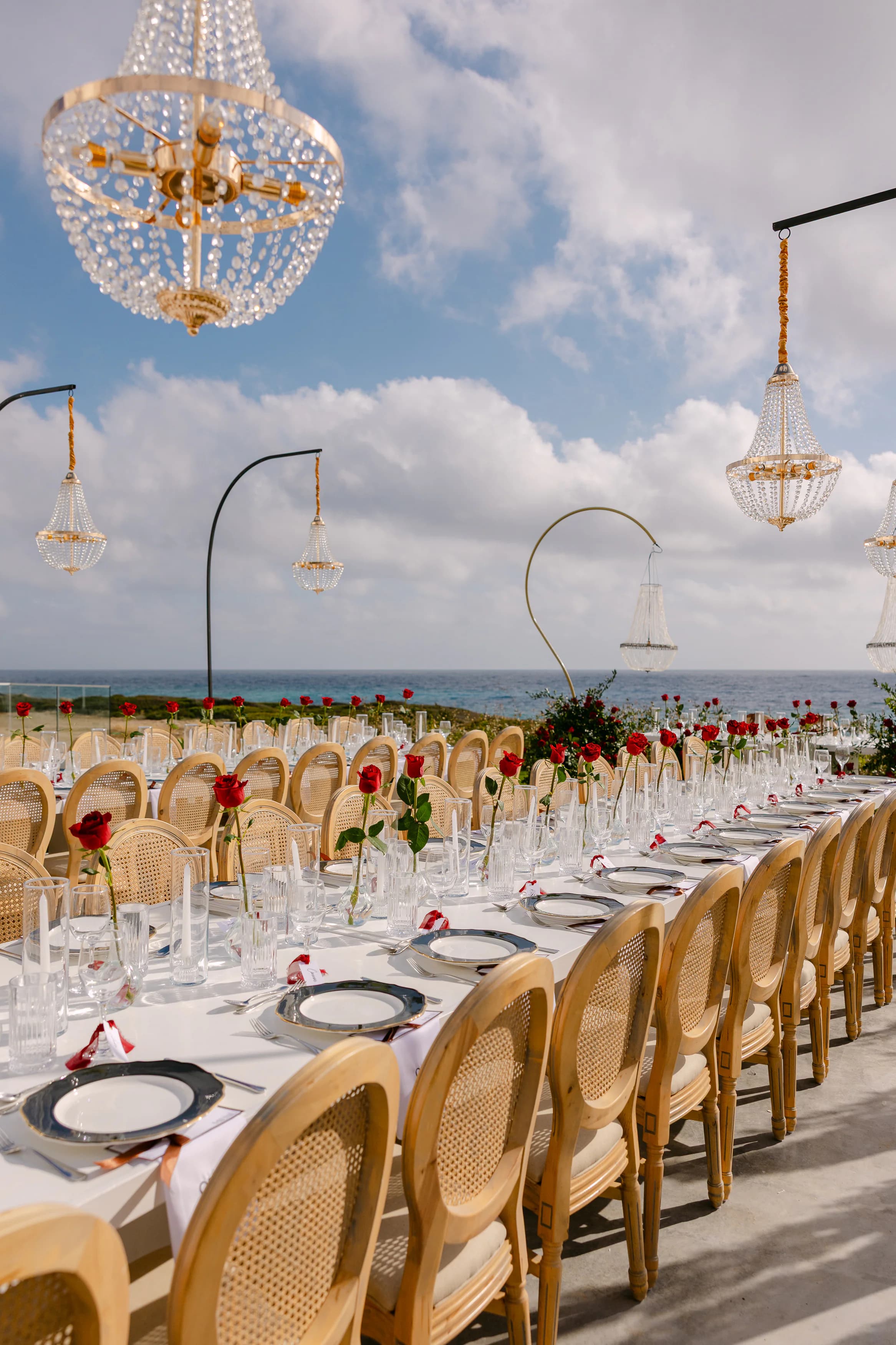 Crystal chandelier over the OZEA wedding banquet table with single red roses, gold arches and the Sea Caves Paphos coastline