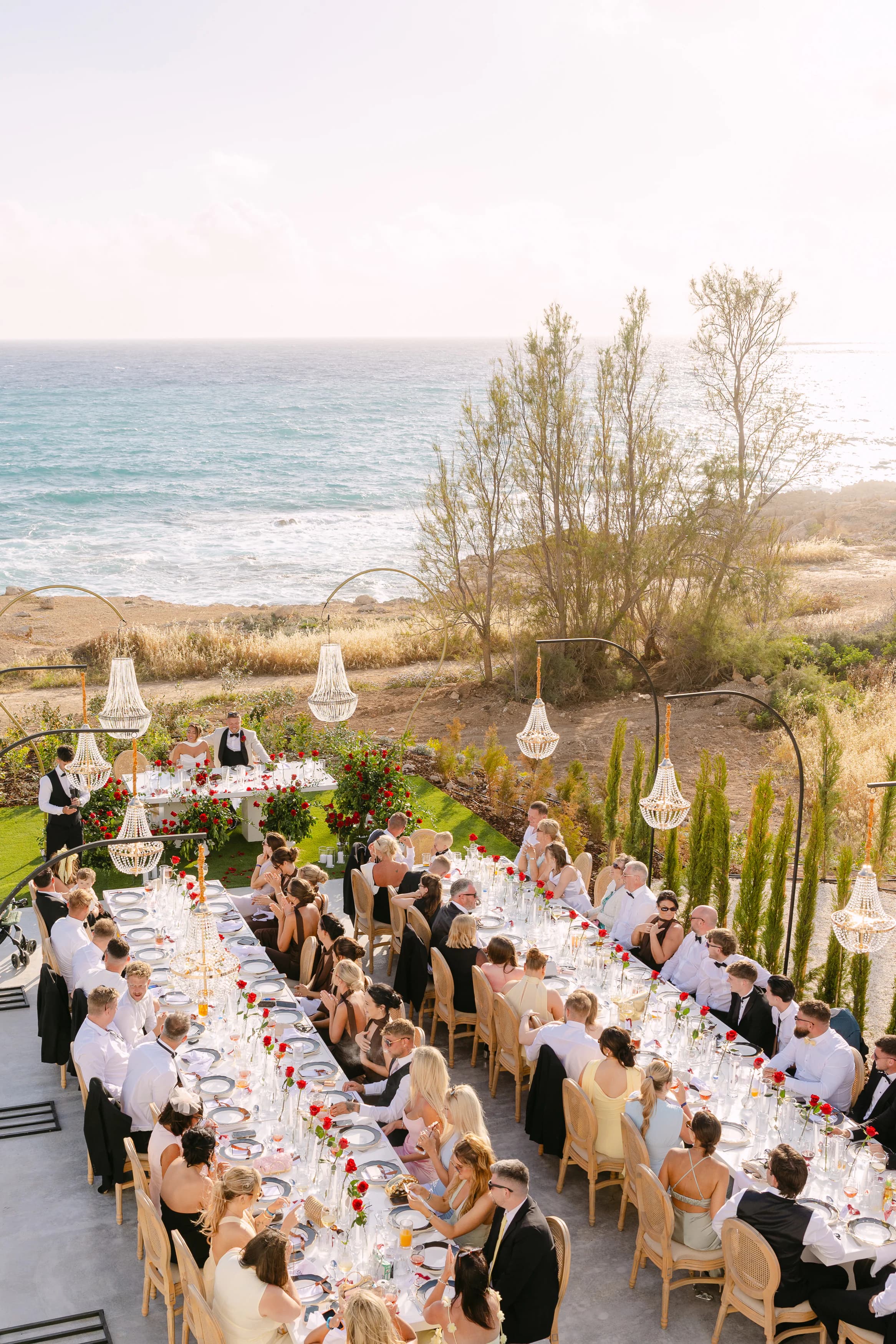 Golden-hour wedding reception at OZEA in Sea Caves Paphos — guests seated along a long banquet table with the Mediterranean Sea behind them