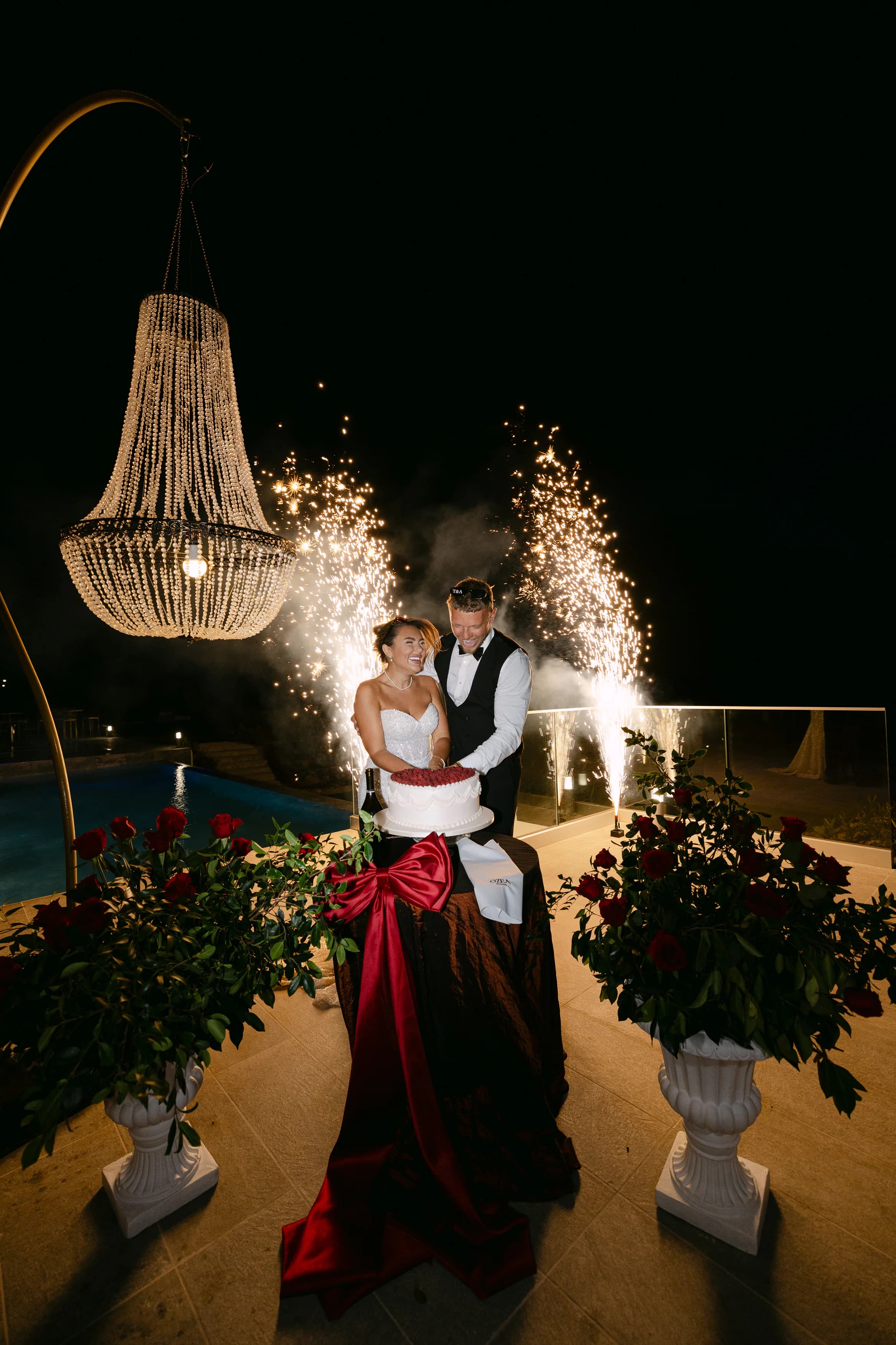 Bride in a red mermaid evening dress and groom cutting the wedding cake at night with sparkler fountains at OZEA Sea Caves Paphos